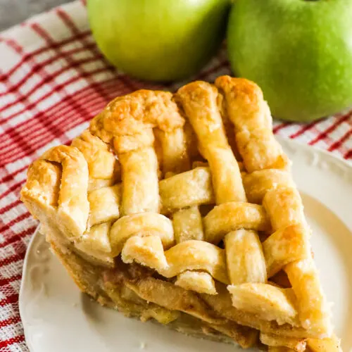 A slice of apple pie with a lattice crust. Two green apples in background.