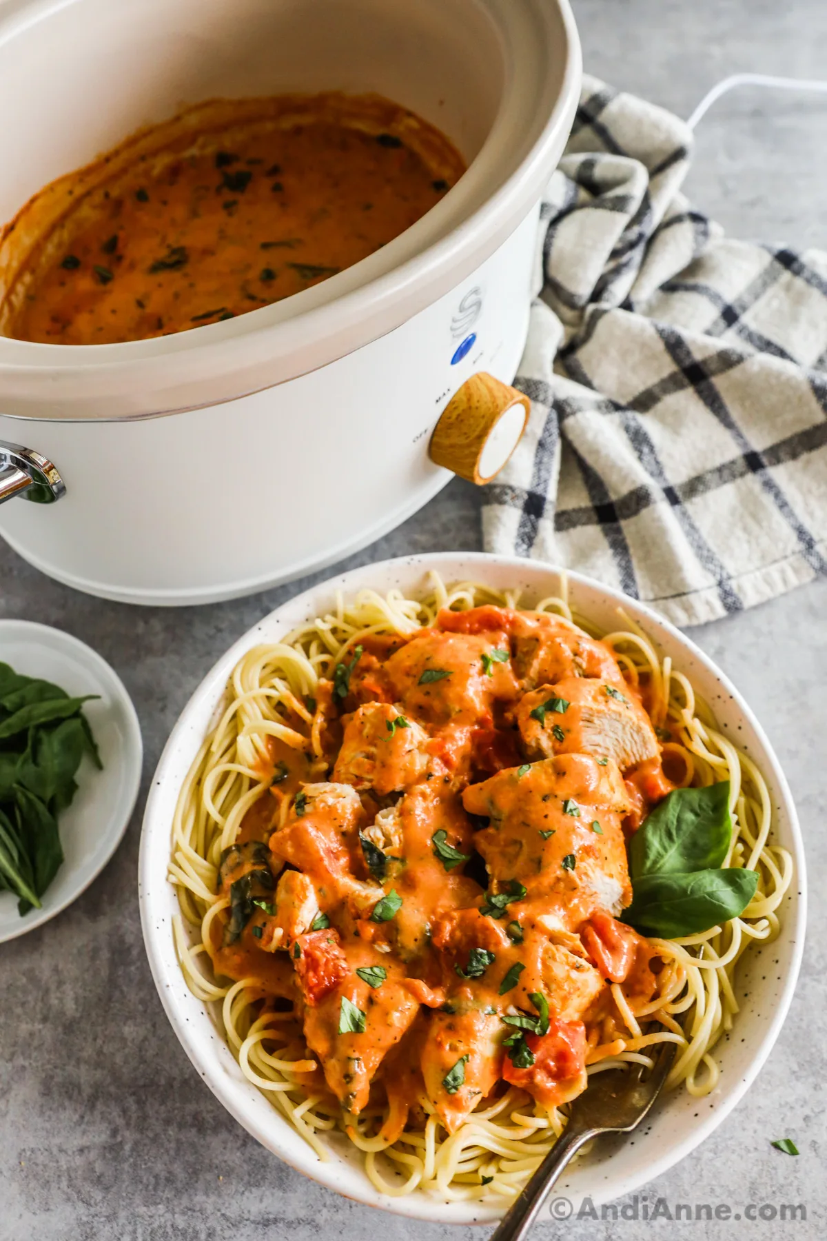 Slow cooker in background with plate of finished recipe in foreground: tomato sauce on sliced chicken breast on top of spaghetti with fresh basil on top.