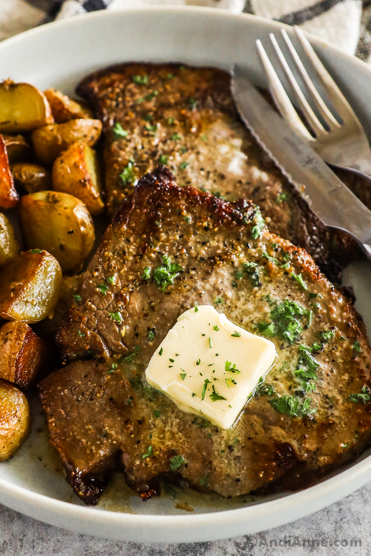 Close up of steaks topped with butter and crispy potatoes on a plate with a fork and knife.