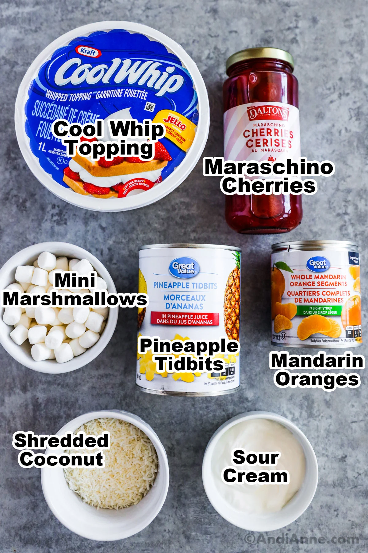 Recipe ingredients on counter including cool whip container, jar of maraschino cherries, mini marshmallows, canned pineapple and mandarin oranges, shredded coconut and sour cream.