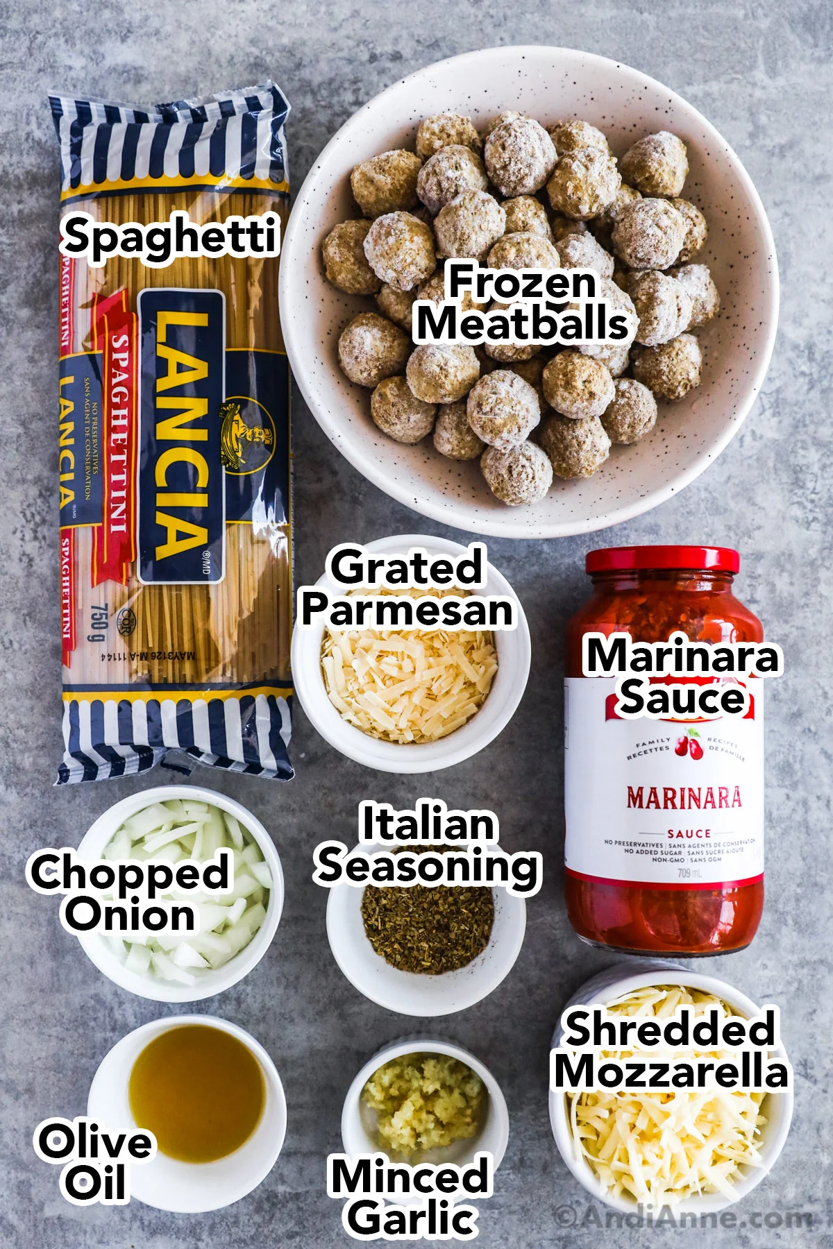Recipe ingredients in bowls including frozen meatballs, packaged spaghetti, jar of marinara, bowls of grated parmesan, shredded mozzarella, chopped onion, Italian seasoning, olive oil, an minced garlic.