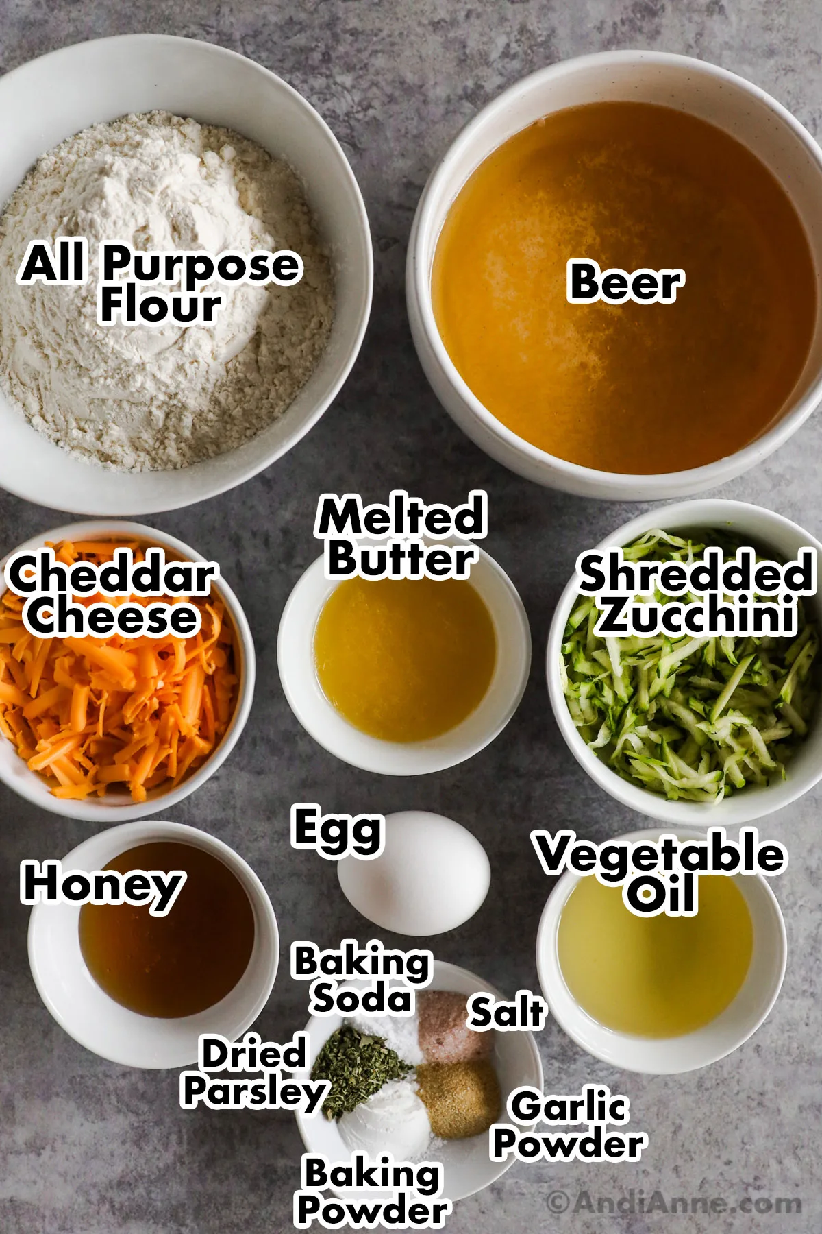 Recipe ingredients in bowls including flour, beer, cheese, butter, shredded zucchini, honey, egg, vegetable oil, dried parsley, garlic powder, baking soda and salt.