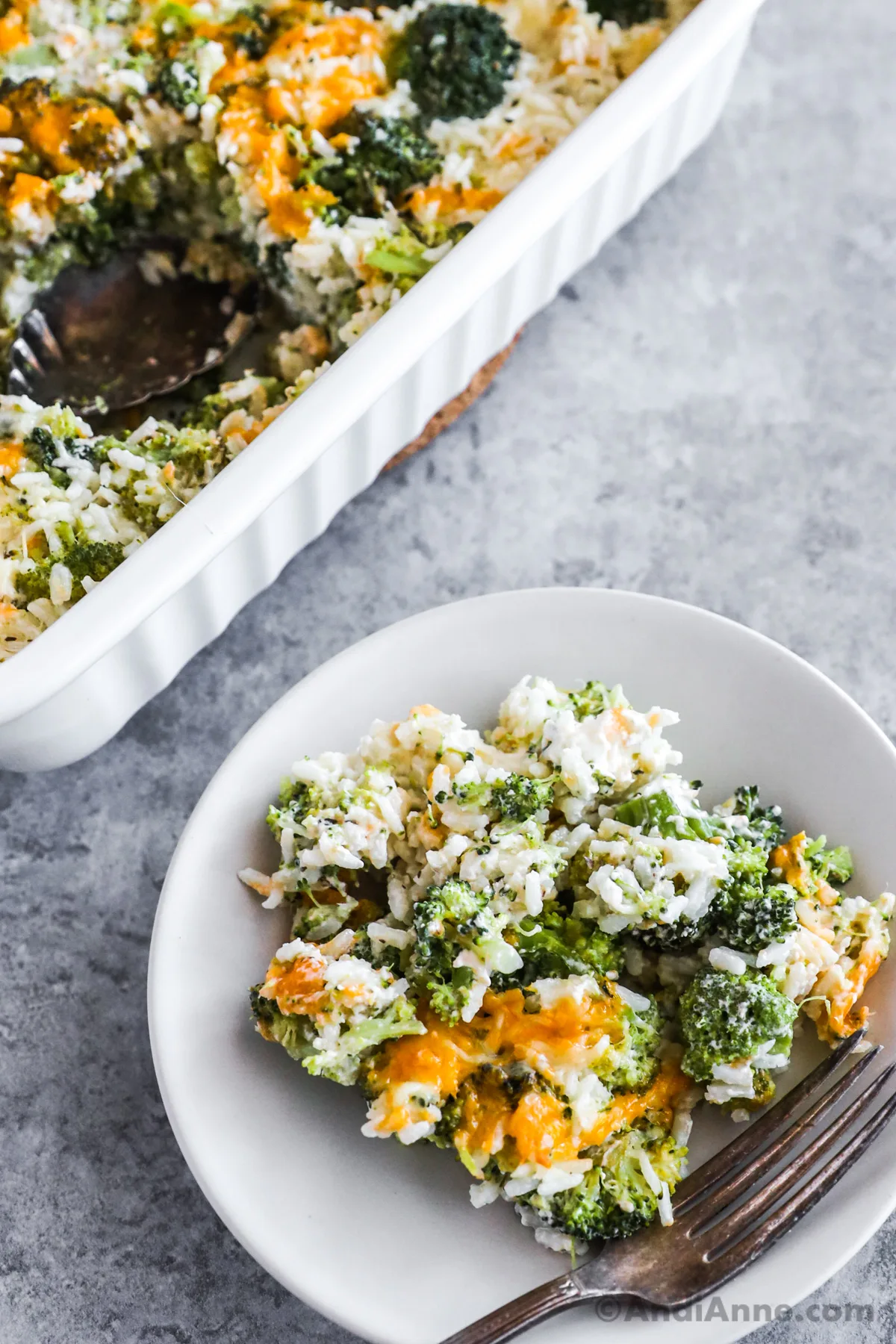 A plate of broccoli cheese rice casserole recipe with a fork, and casserole dish behind it.