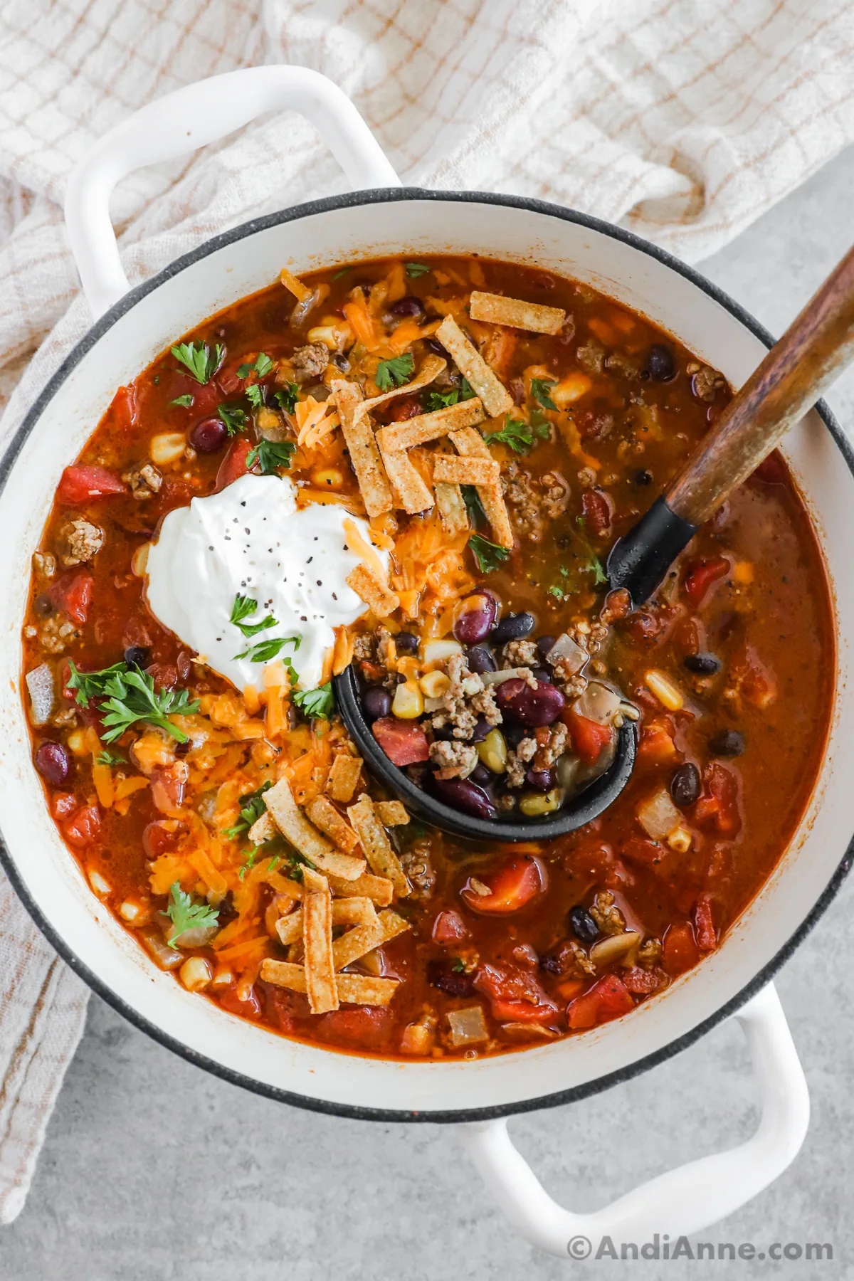 A bowl of ground beef taco soup with a dollop of sour cream and tortilla strips and a soup ladle.