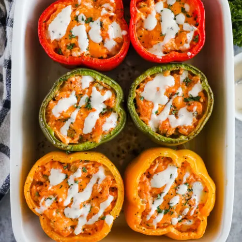 Six roasted bell peppers stuffed with buffalo chicken and drizzled with ranch dressing all in a casserole dish.