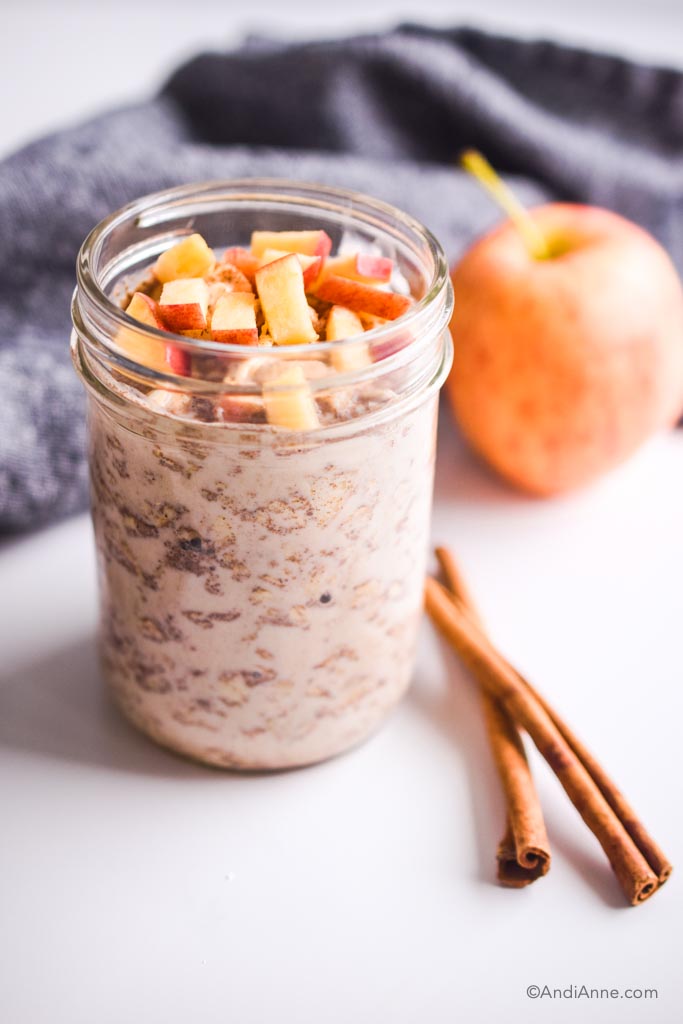 Image of apple cinnamon overnight oats in a small mason jar. Sliced apples on top and cinnamon sticks on the table. 