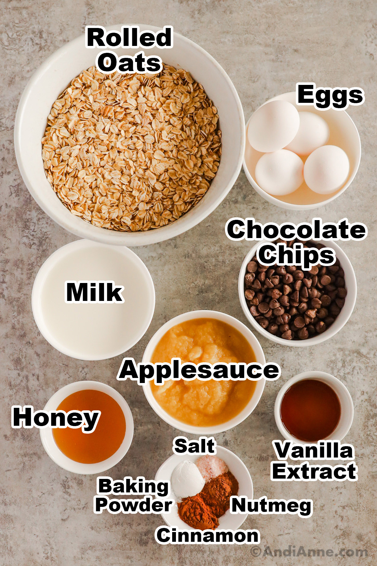 Recipe ingredients in bowls including rolled oats, eggs, milk, chocolate chips, applesauce, honey, vanilla and spices.