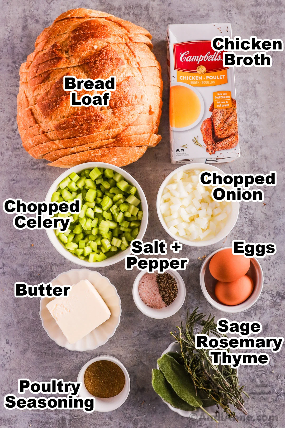 Recipe ingredients on a counter including a loaf of bread, carton of broth, bowls of chopped celery, chopped onion, butter, eggs, salt and pepper, poultry seasoning, and fresh herbs.