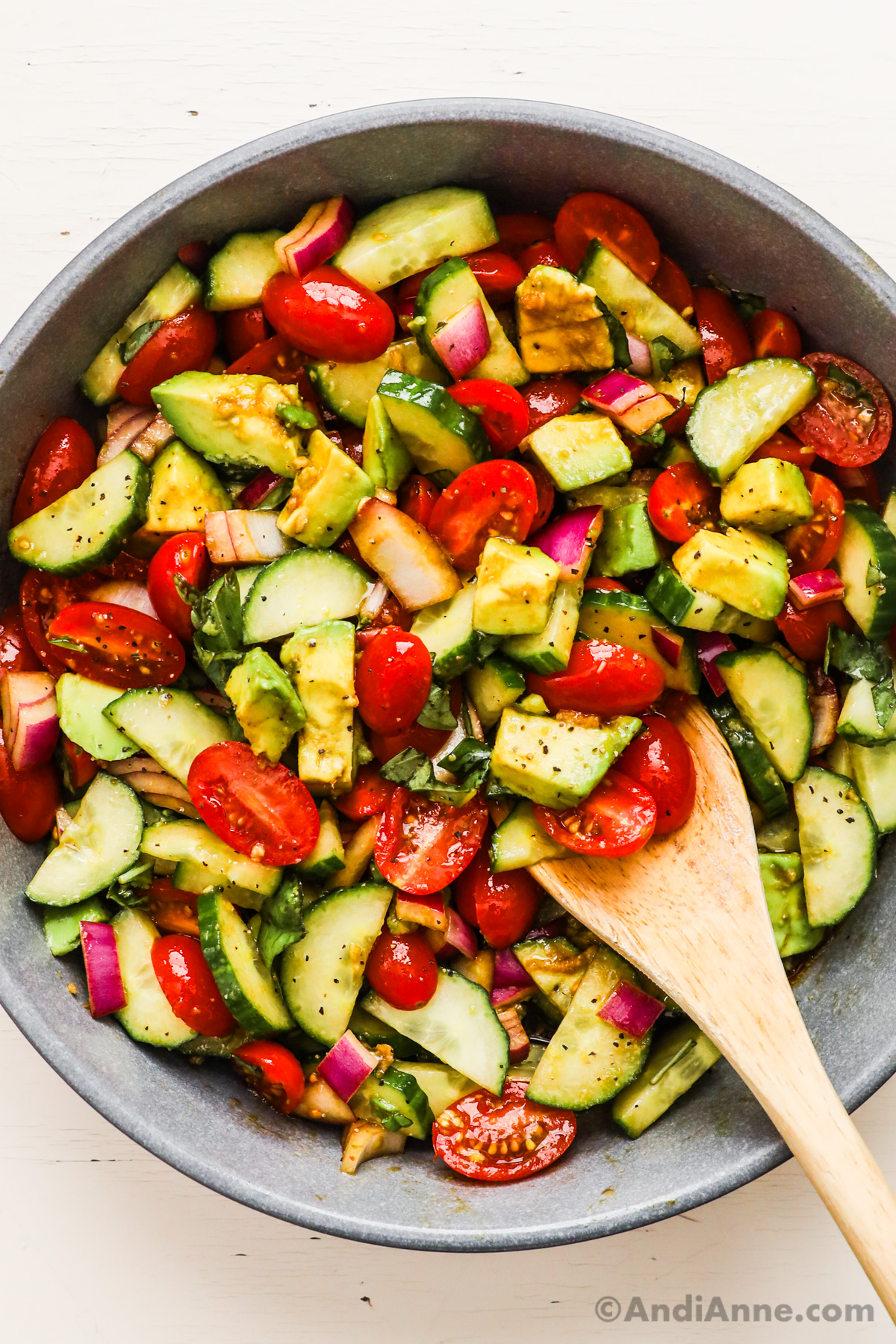 Close up of chopped avocado, cucumber, tomato, red onion and basil salad with a wood spoon.