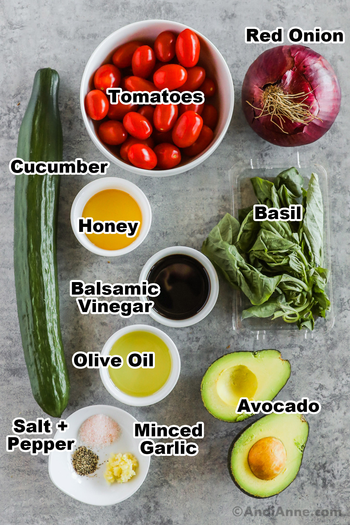 Recipe ingredients including bowl of grape tomatoes, a red onion, an english cucumber, a bunch of fresh basil, a sliced avocado, bowls of honey, balsamic vinegar, olive oil, and a plate with salt and pepper and minced garlic.