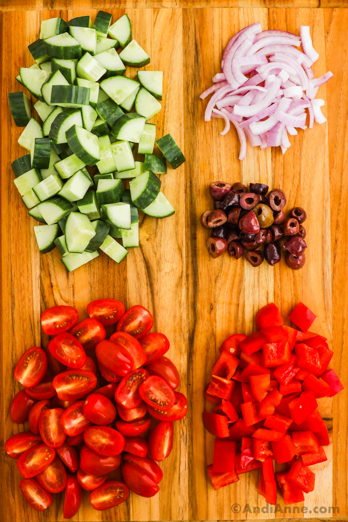 Chopped cucumbers, red onion, olives, tomatoes, and red peppers on a cutting board.