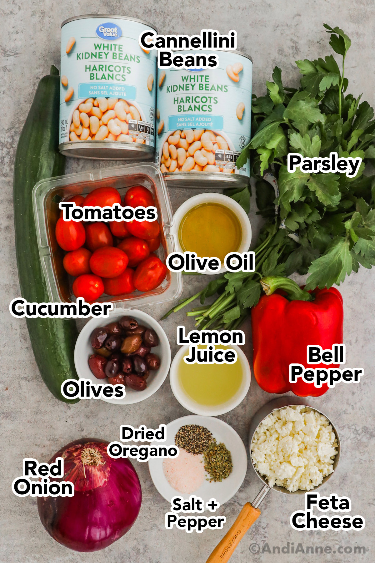 Recipe ingredients on counter including cans of cannellini beans, parsley, cucumber, tomatoes, bell pepper, red onion and bowls of olives, lemon juice, feta cheese, salt and pepper