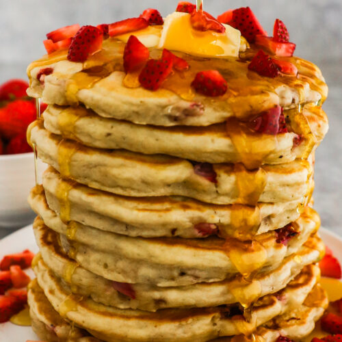 A stack of strawberry pancakes topped with butter, chopped strawberries and drizzled with maple syrup