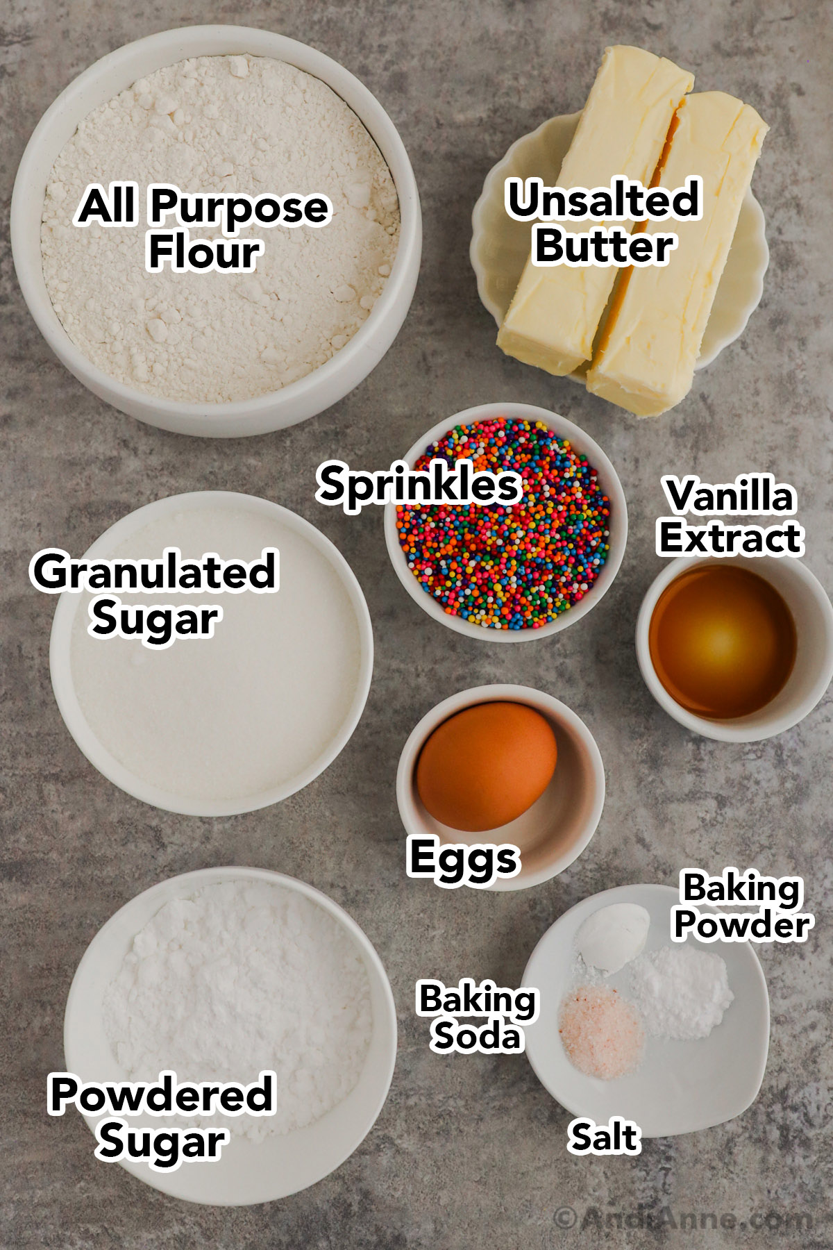 Recipe ingredients in bowls including bowl of flour, butter sticks, granulated sugar, sprinkles, vanilla, eggs, and powdered sugar.