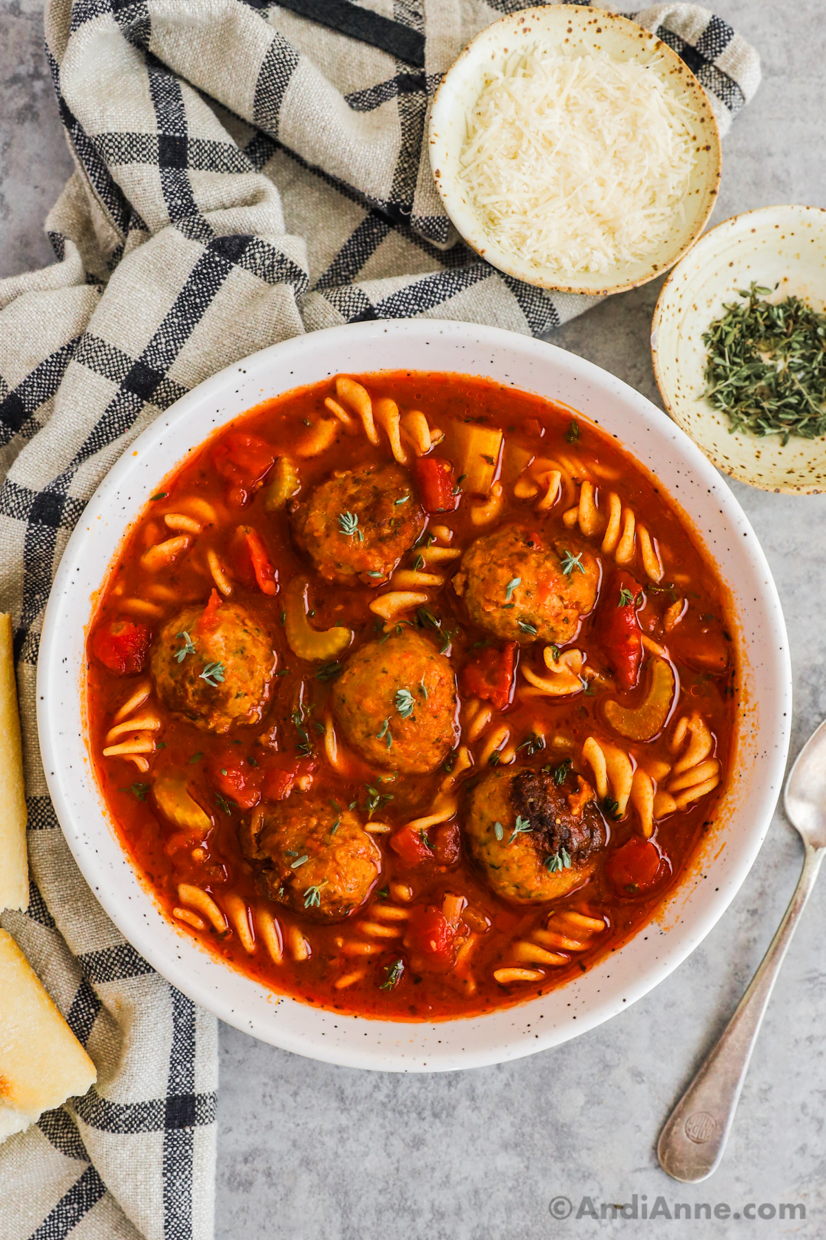 Bowl of italian meatball soup with spoon, and small bowls of ingredients surrounding it