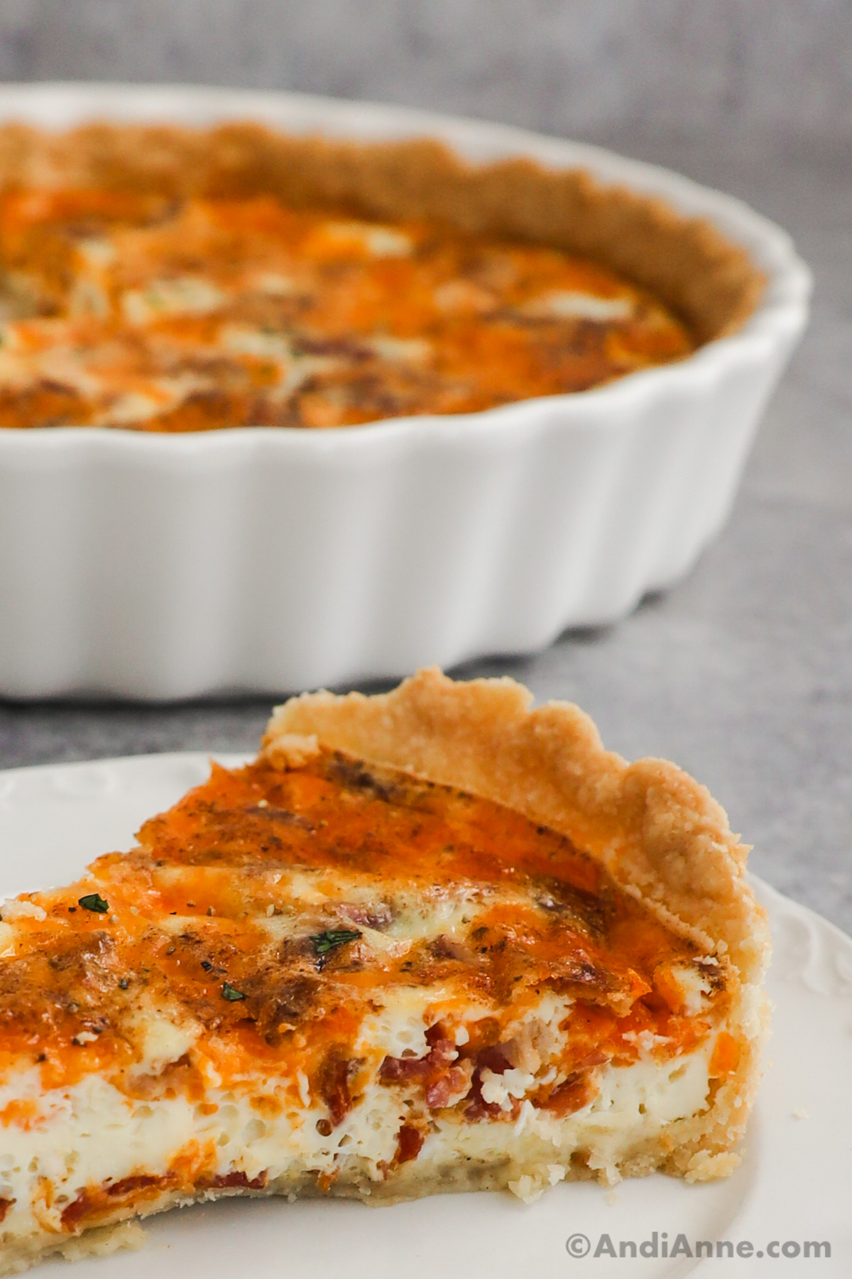 Close up of a slice of bacon cheese quiche with the quiche pan and the rest of the quiche in the background.