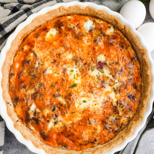 A bacon and cheese quiche in pie plate with forks, eggs and kitchen towel surrounding it