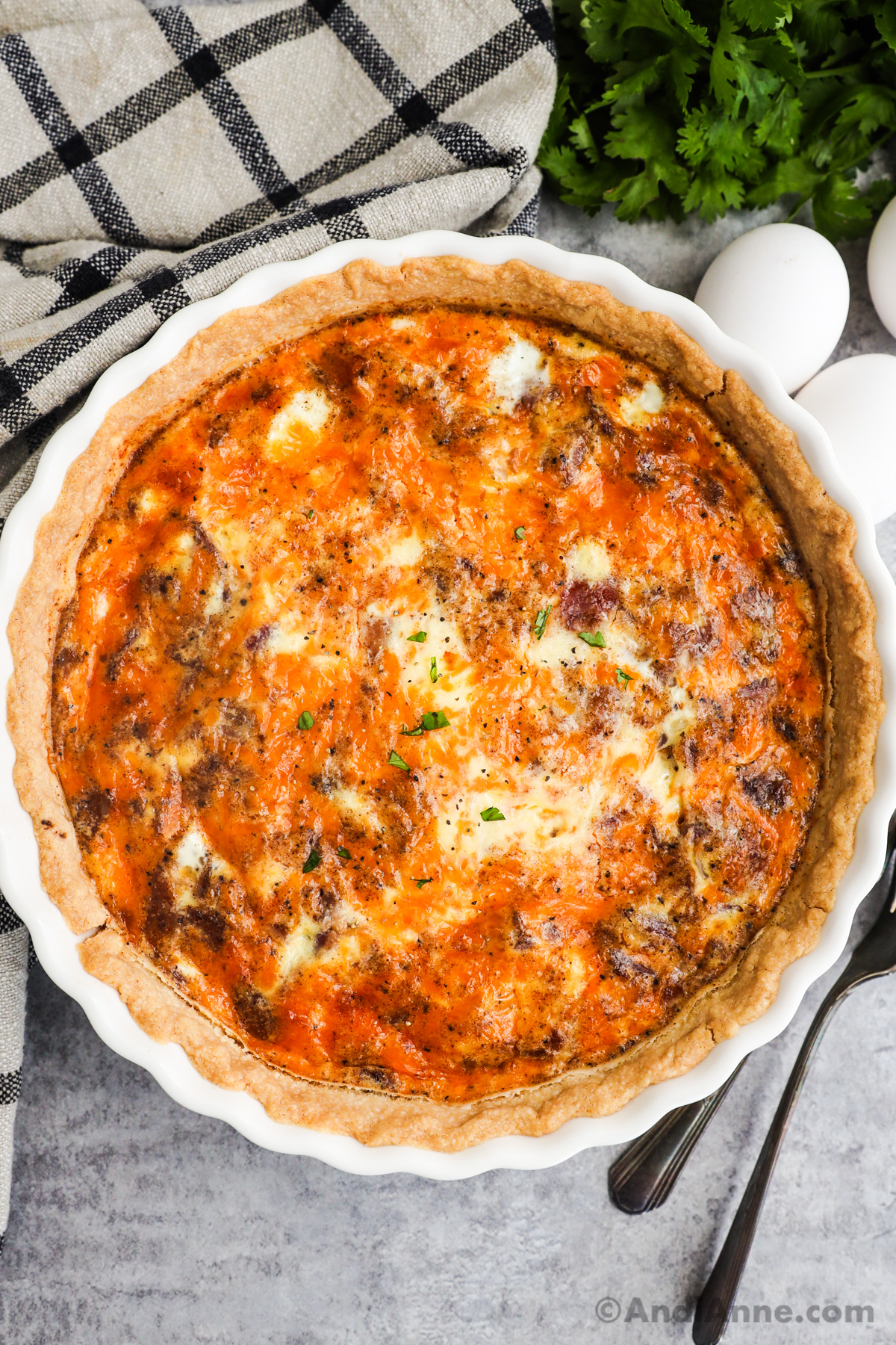 Photo of a bacon cheese quiche in a white quiche dish with eggs and kitchen towel surrounding it.