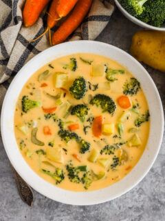 A bowl of cheddar broccoli potato soup with carrots and potatoes surrounding it