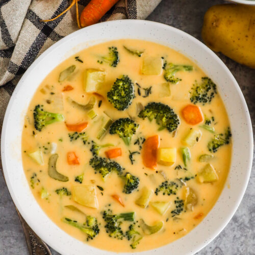 A bowl of cheddar broccoli potato soup with carrots and potatoes surrounding it