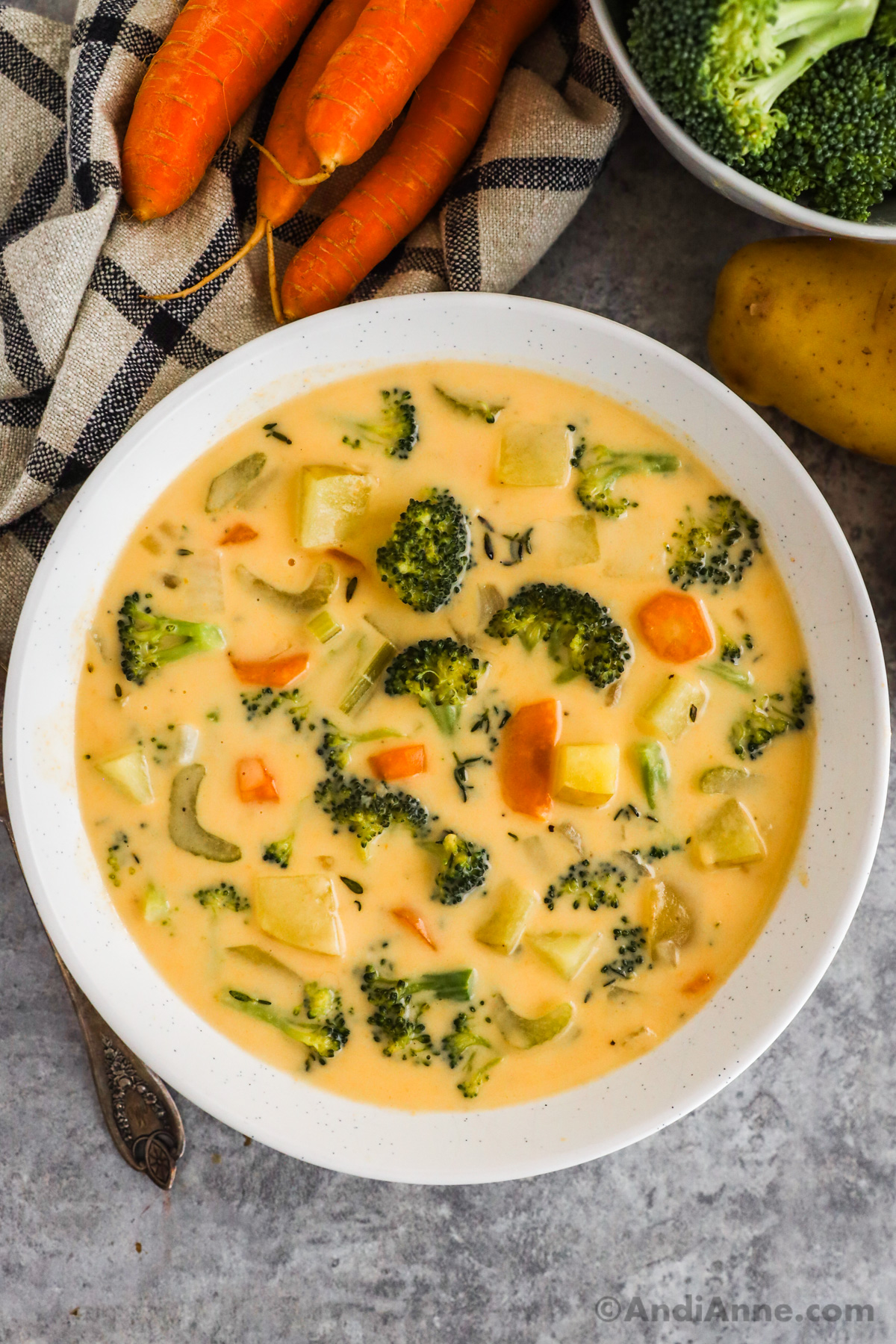 A bowl of broccoli cheddar potato soup with carrots, potato and broccoli around it.