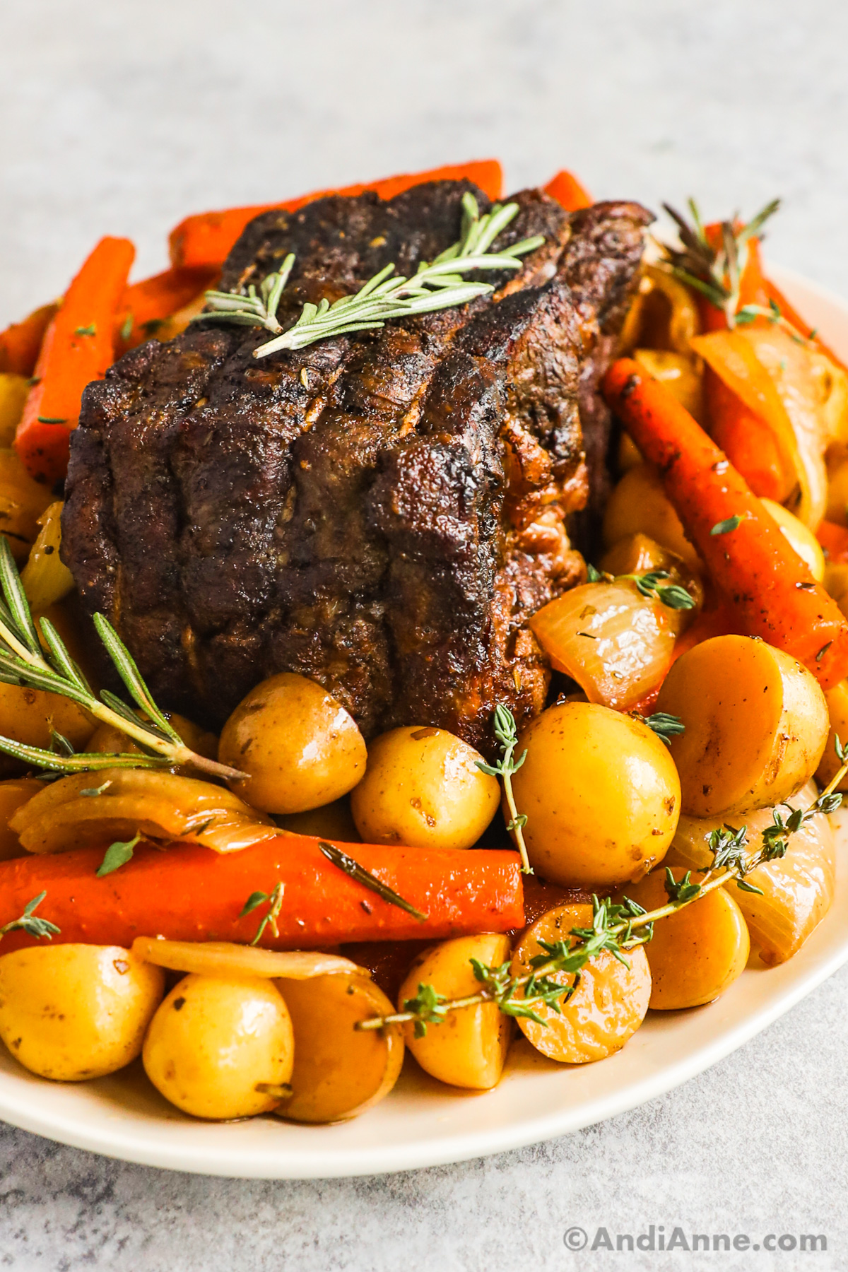 Close up of cooked pot roast surrounded by carrots, potatoes and onion chunks, topped with sprigs of rosemary and thyme.