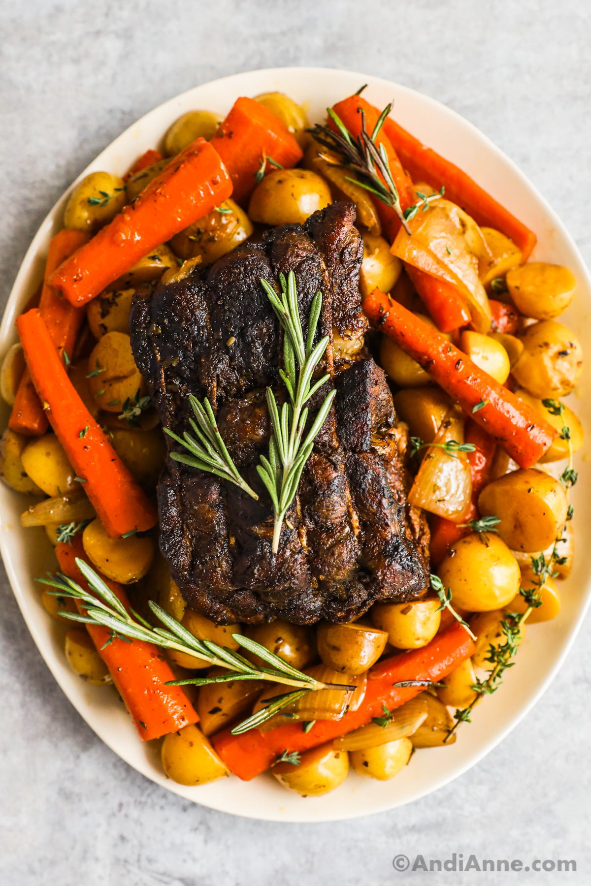 A plate with cooked beef surrounded by potatoes and carrots and fresh sprigs of thyme and parsley.