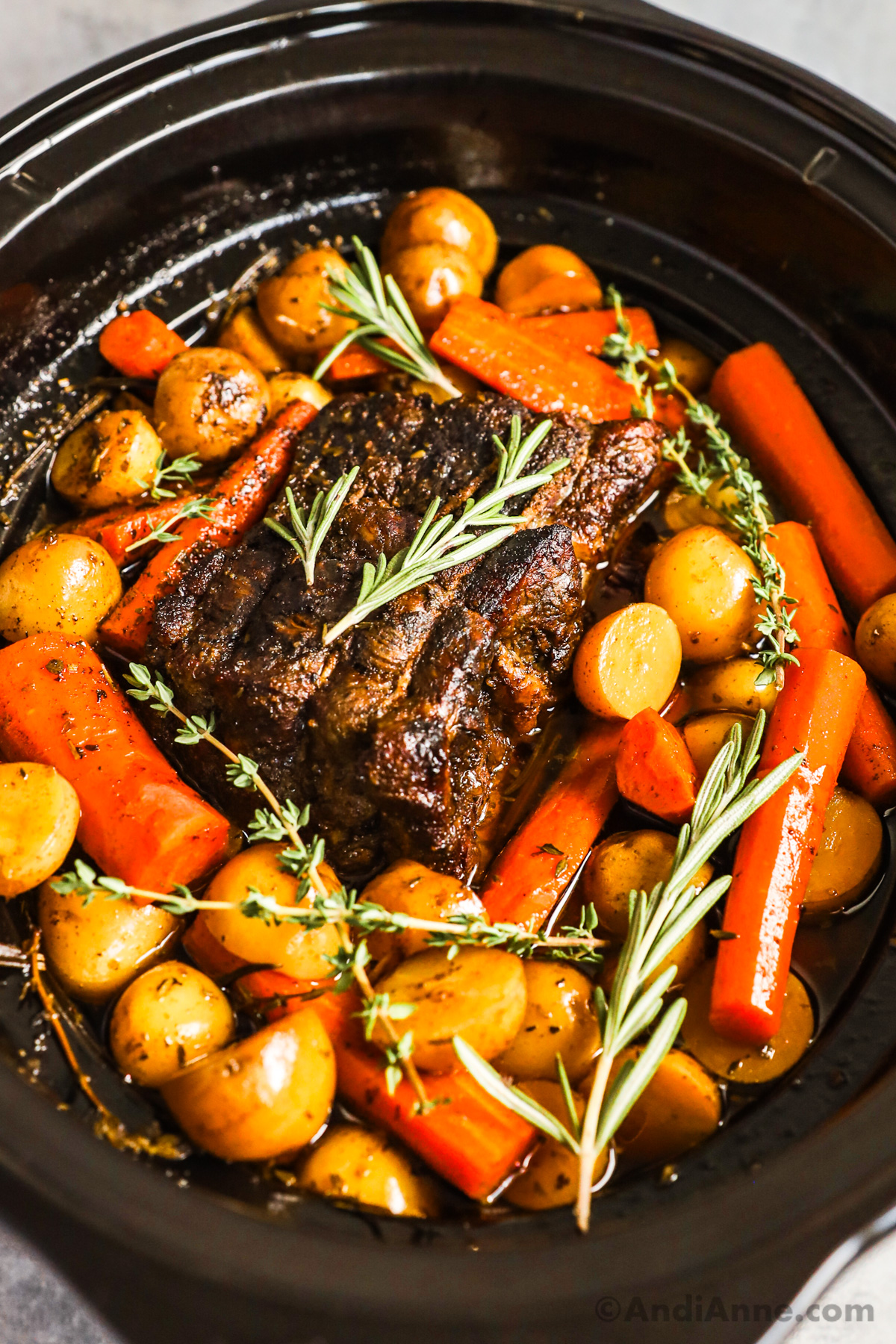 A slow cooker with a pot roast, surrounded by carrot chunks, potatoes and fresh herbs.