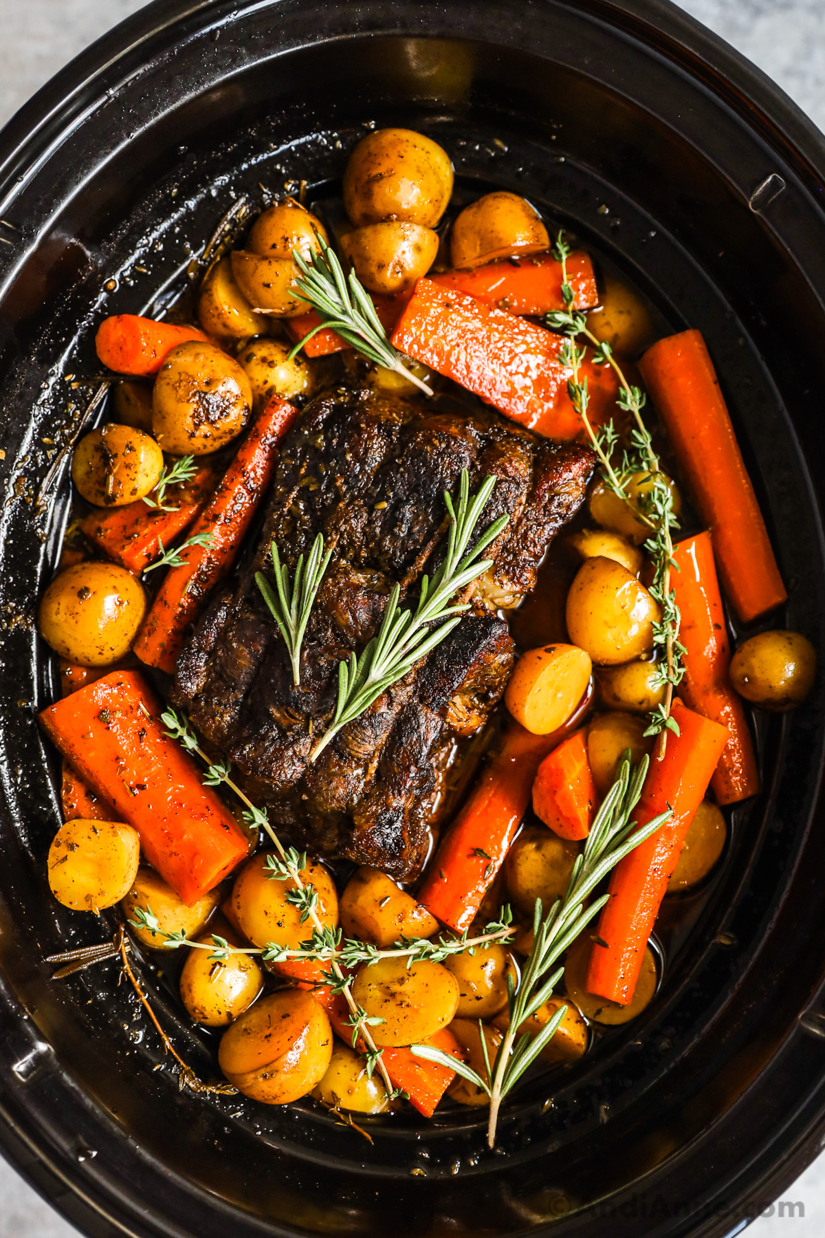 Looking into a slow cooker with a beef roast, carrots, tomatoes, sprigs of rosemary and thyme