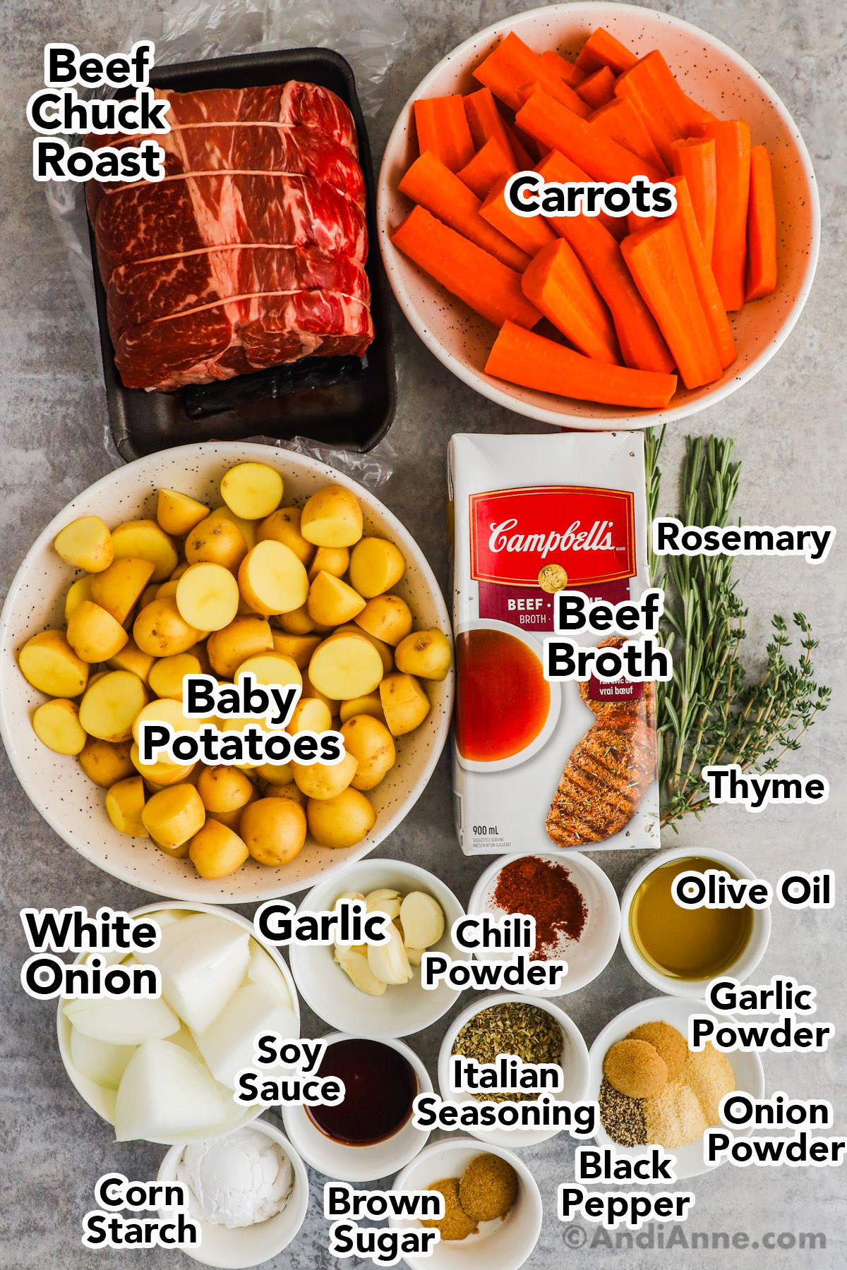 Recipe ingredients including a raw beef chuck roast, bowls with chopped carrots, chopped potatoes, carton of beef broth, sprigs of fresh rosemary and thyme, chopped white onion, garlic cloves, spices, and olive oil.