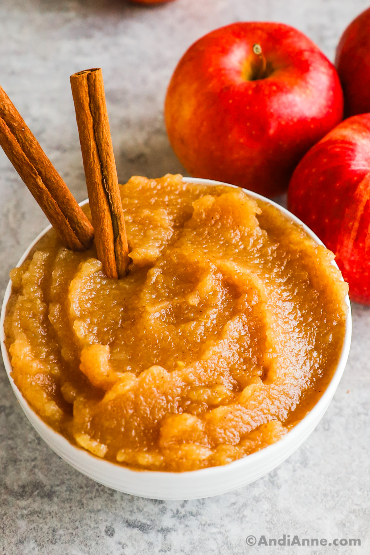 Bowl of applesauce with two cinnamon sticks poking out. Some fresh apples are behind the bowl.