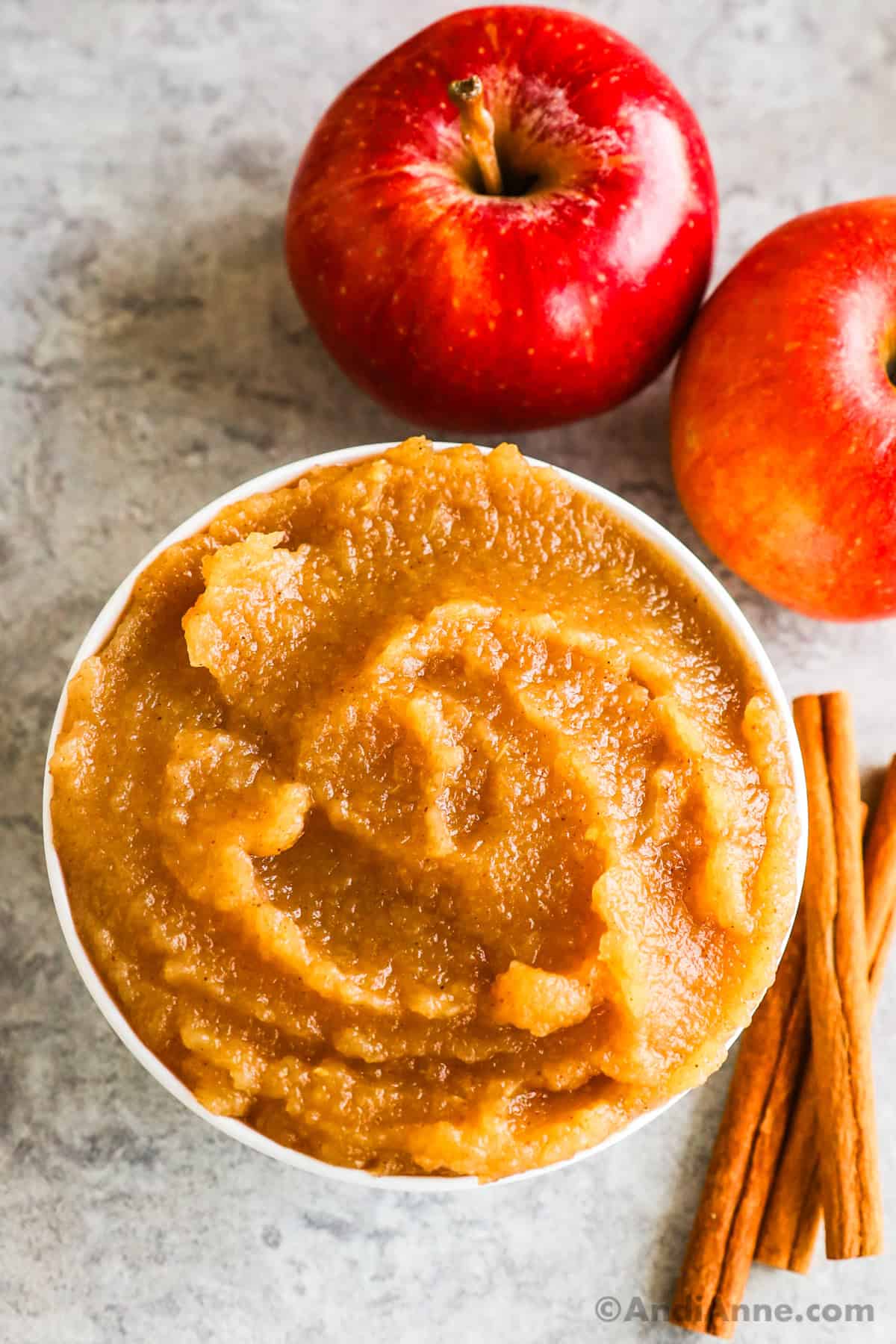 A bowl of homemade applesauce with cinnamon sticks and apples surrounding the bowl.