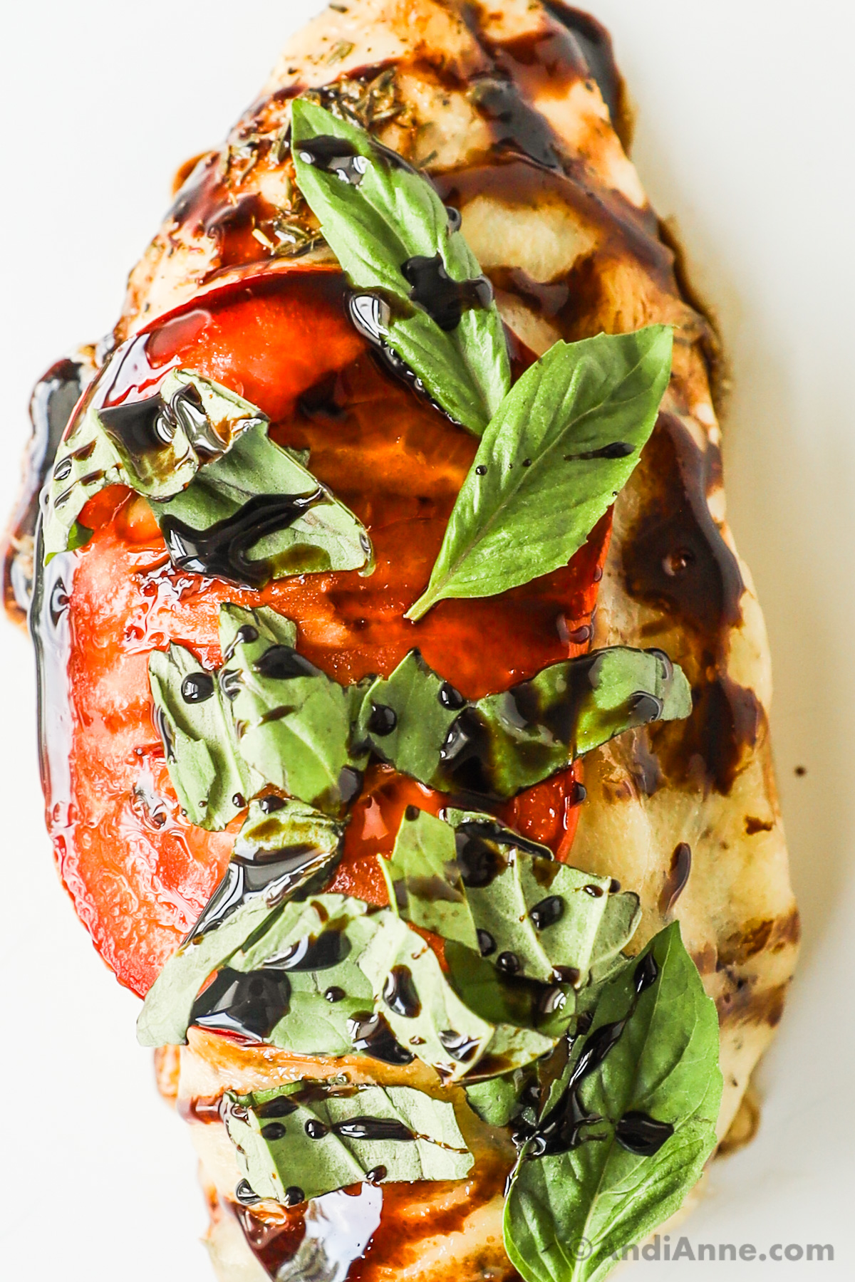 Close up of a chicken breast topped with melted cheese, tomato slice, fresh basil and drizzled with balsamic vinegar.