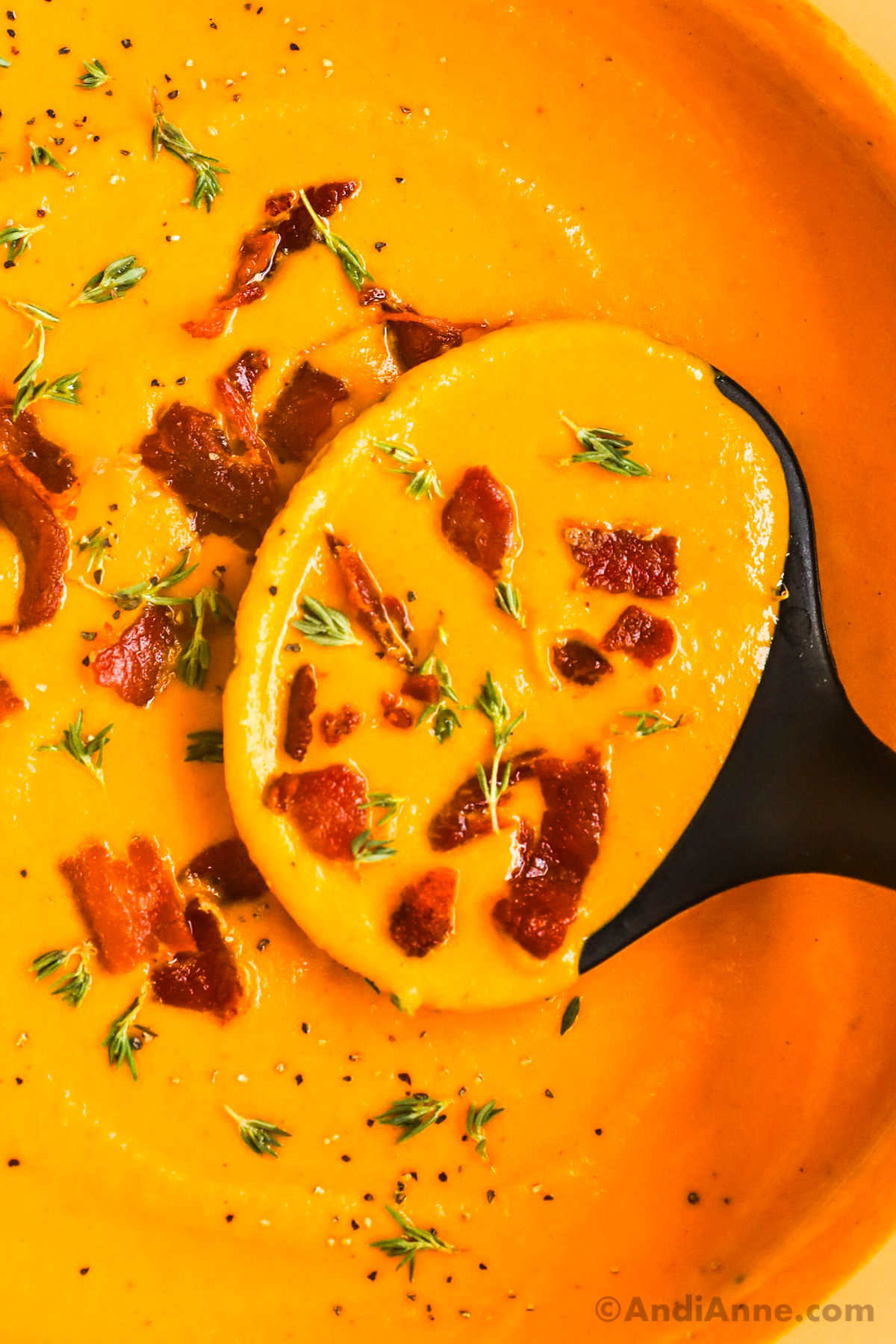 Close up of soup ladle scooping creamy carrot soup with bacon and thyme pieces.