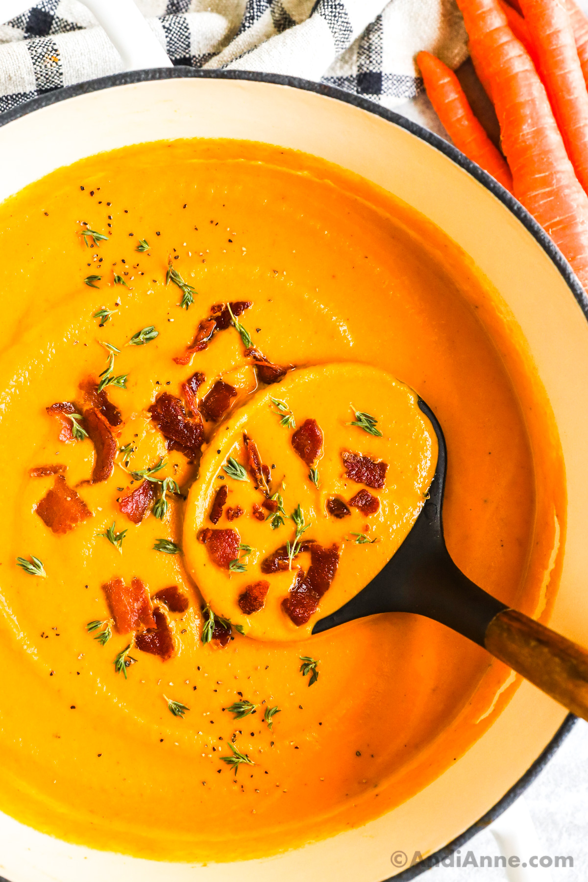 A soup ladle in creamy carrot soup topped with crumbled bacon, fresh thyme and black pepper.