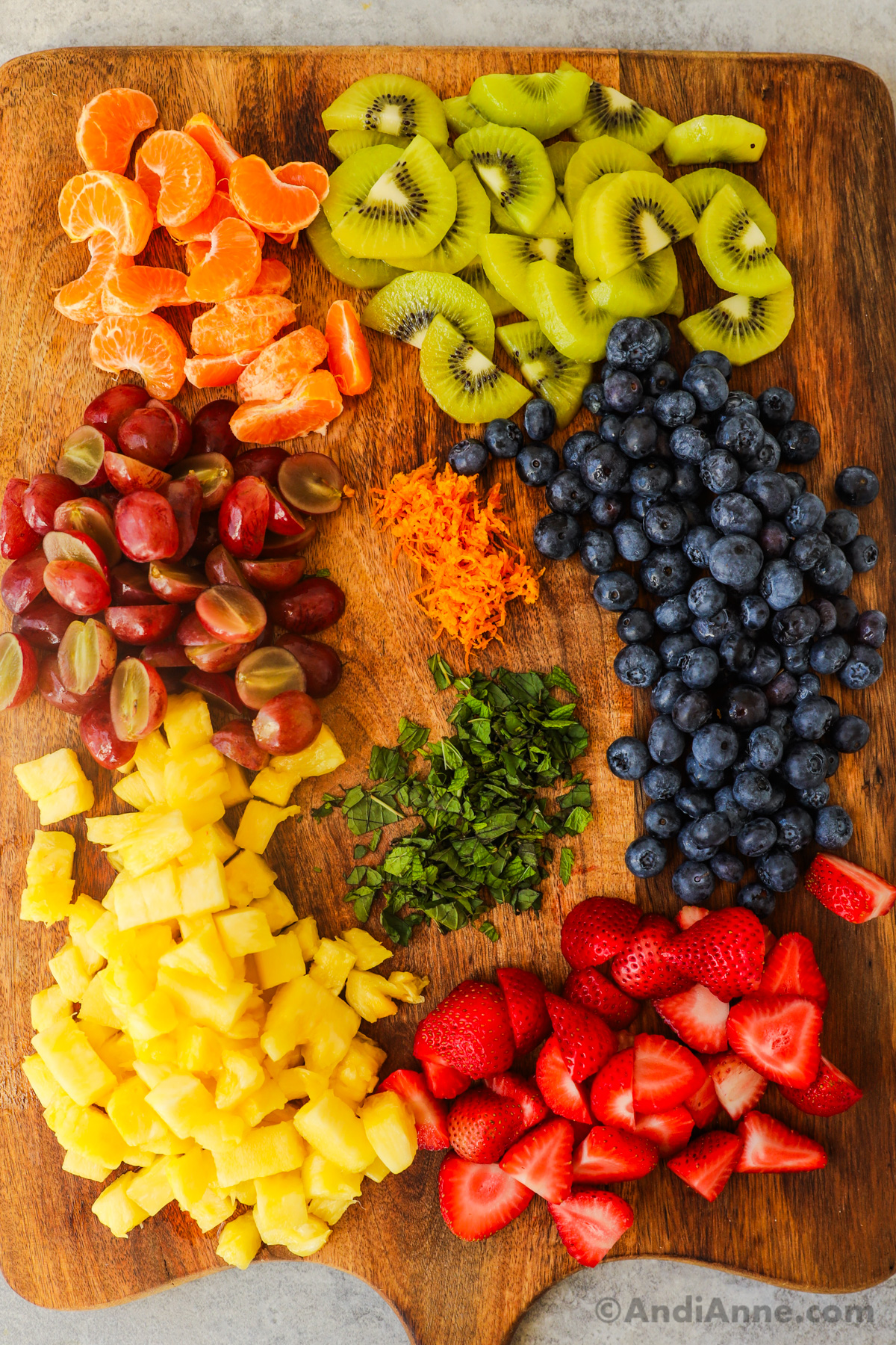 Sliced kiwi, grapes, pineapple, strawberries, chopped mint, orange zest, mandarin orange slices and blueberries all grouped on a cutting board.