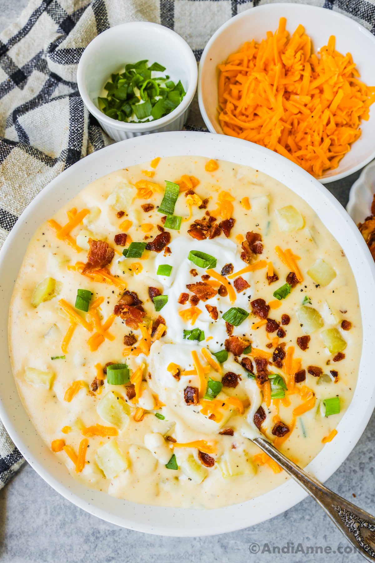 A bowl of potato soup topped with sour cream, crumbled bacon, shredded cheese and chopped scallions. 