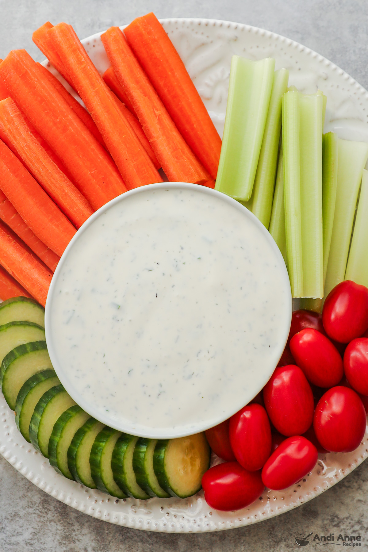 A bowl of ranch dressing with chopped carrots, celery, tomatoes and cucumber surrounding it.