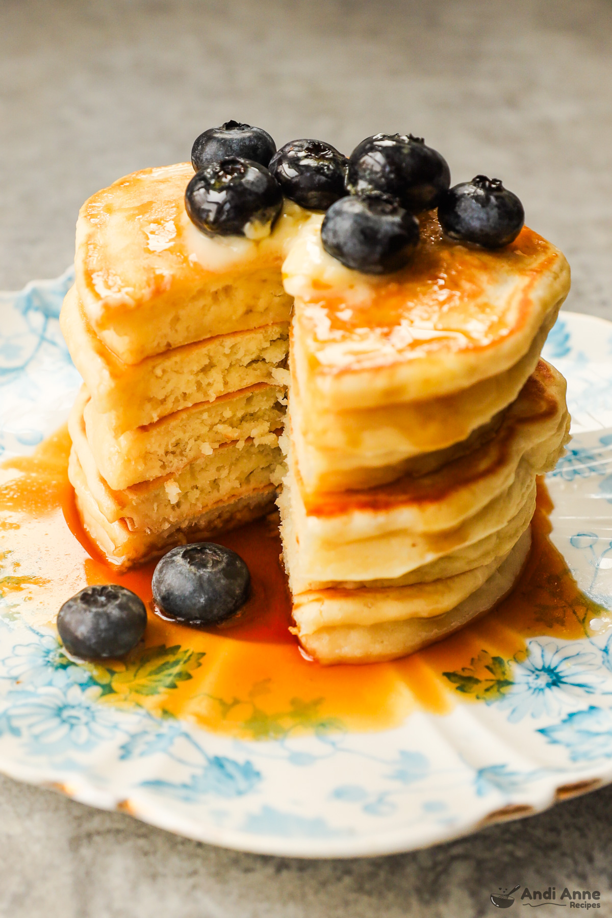 Stack of greek yogurt pancakes with pie shaped slice cut out, drizzled with maple syrup topped with blueberries.