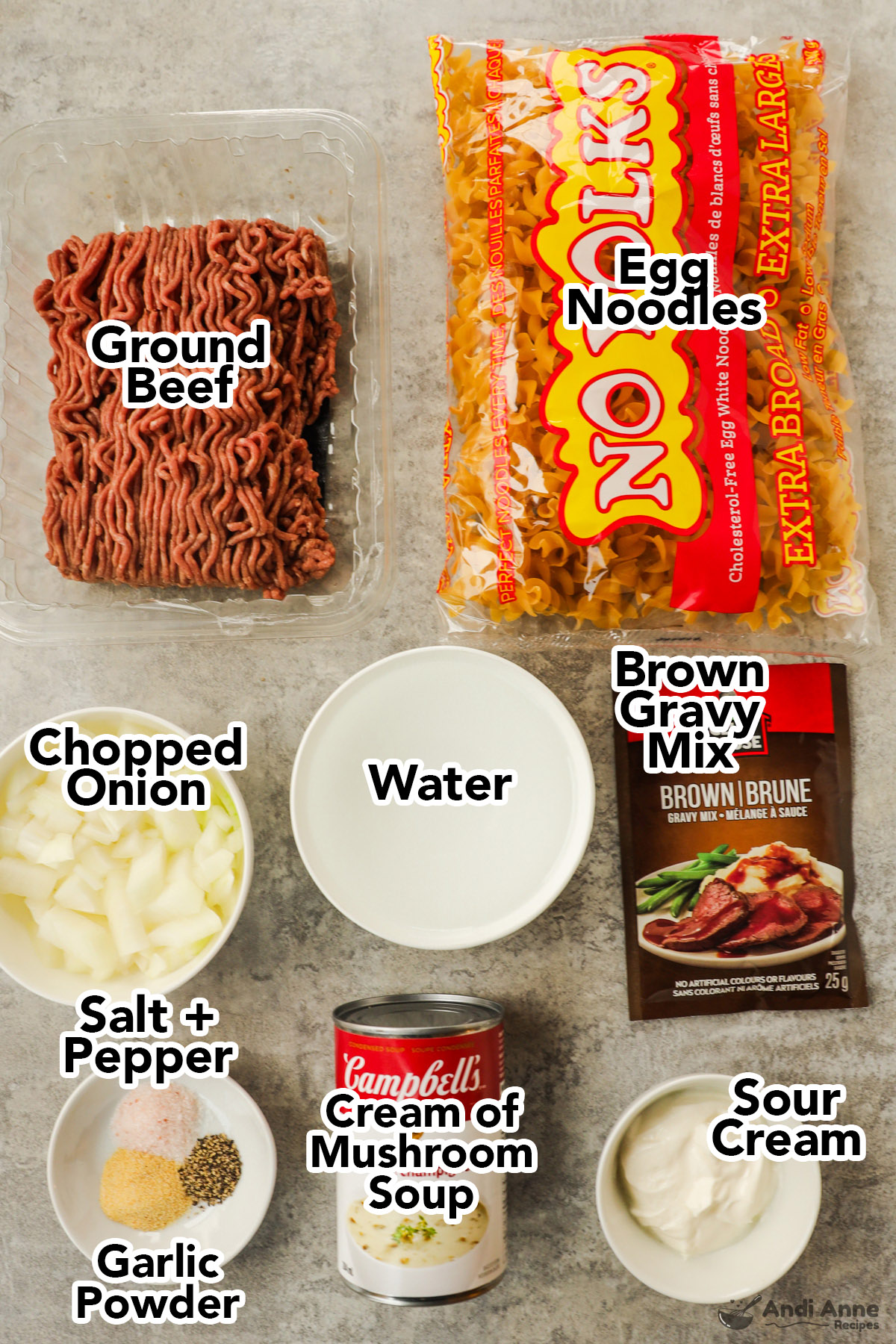 Recipe ingredients including raw ground beef, bag of egg noodles, bowls of chopped onion, water, packet of brown gravy mix, can of mushroom soup, bowl of sour cream, and spices.