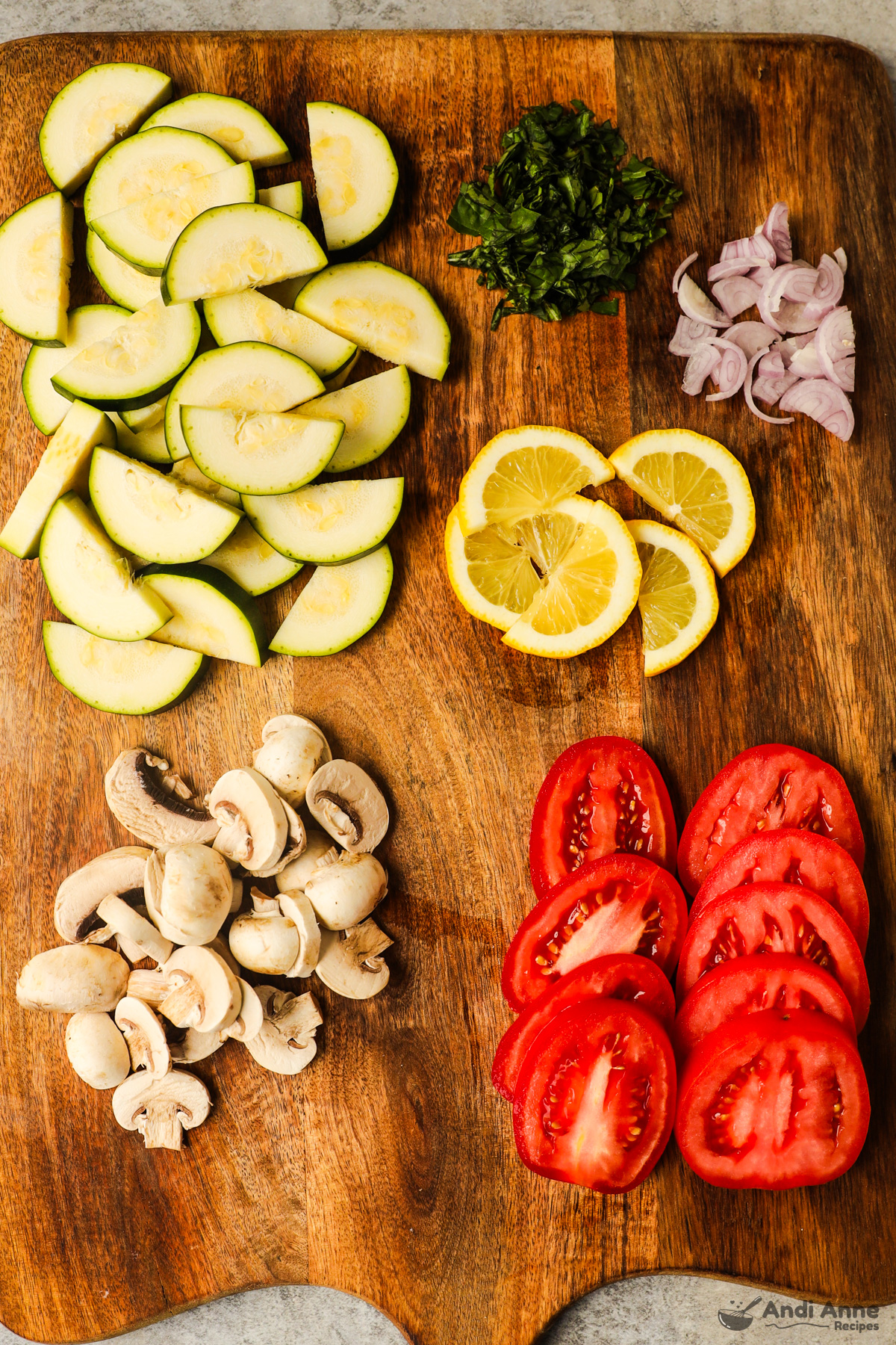 Sliced zucchini, basil, lemon, shallot, mushrooms, and tomatoes on a cutting board.