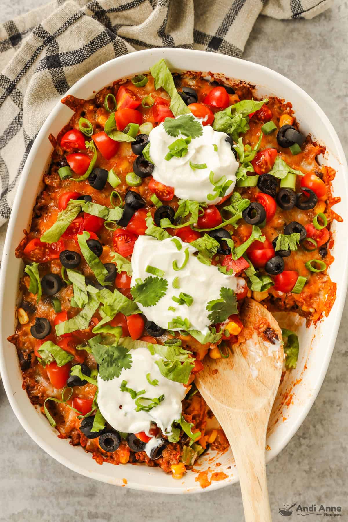 White casserole dish with taco casserole recipe inside with lots of fresh toppings like lettuce, olives, tomatoes and dollops of sour cream sprinkled over top