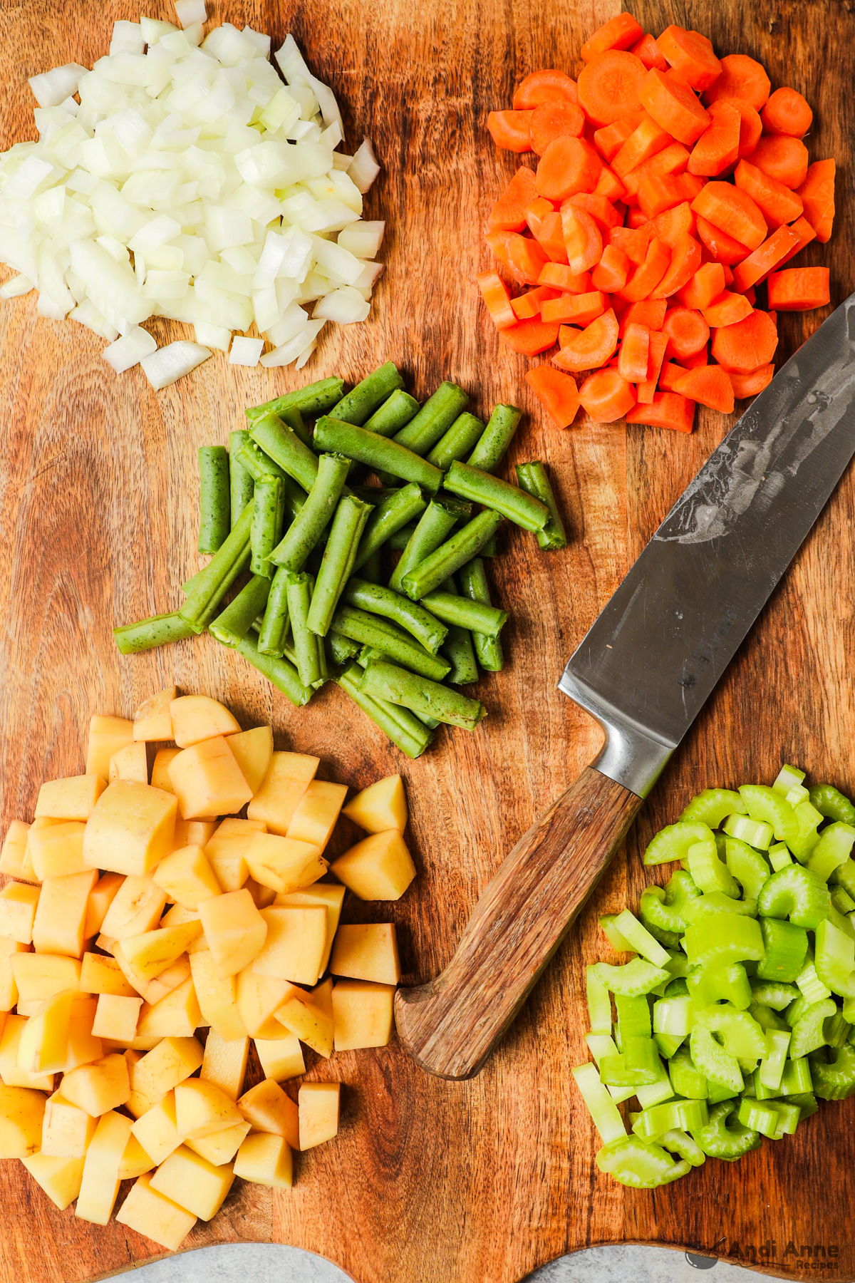 Chopped onion, carrots, green beans, potatoes and celery on a cutting board with a knife.