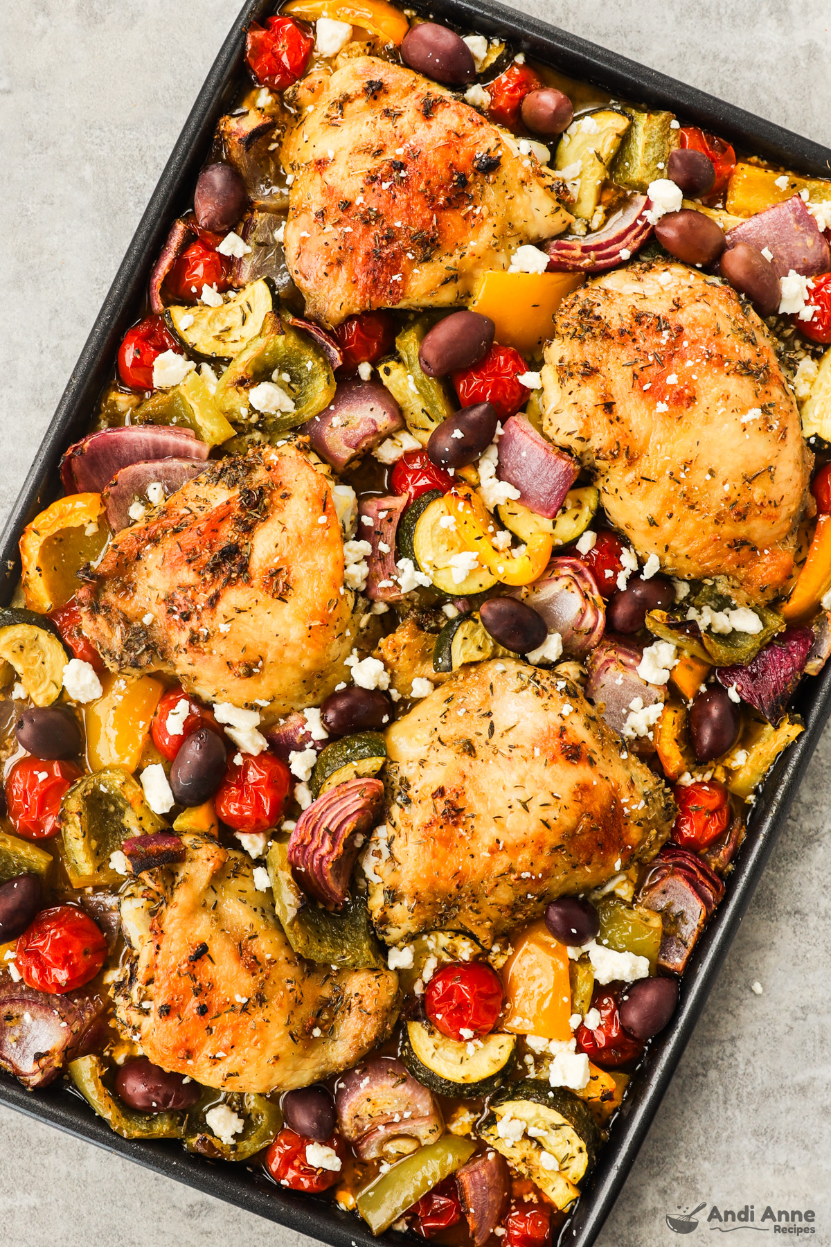 Close up of cooked chicken thighs and veggies on a baking sheet sprinkled with feta cheese.