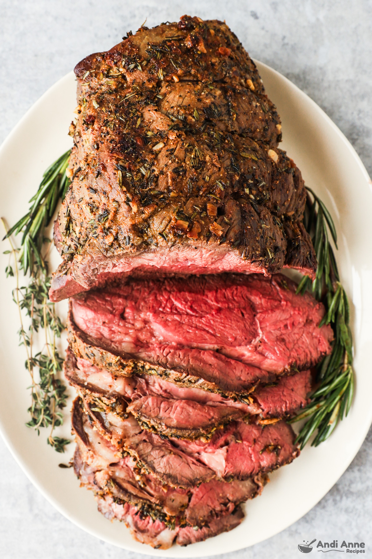 Looking down at a sirloin tip roast with half cut into slices, all on a serving plate with fresh rosemary surrounding it.