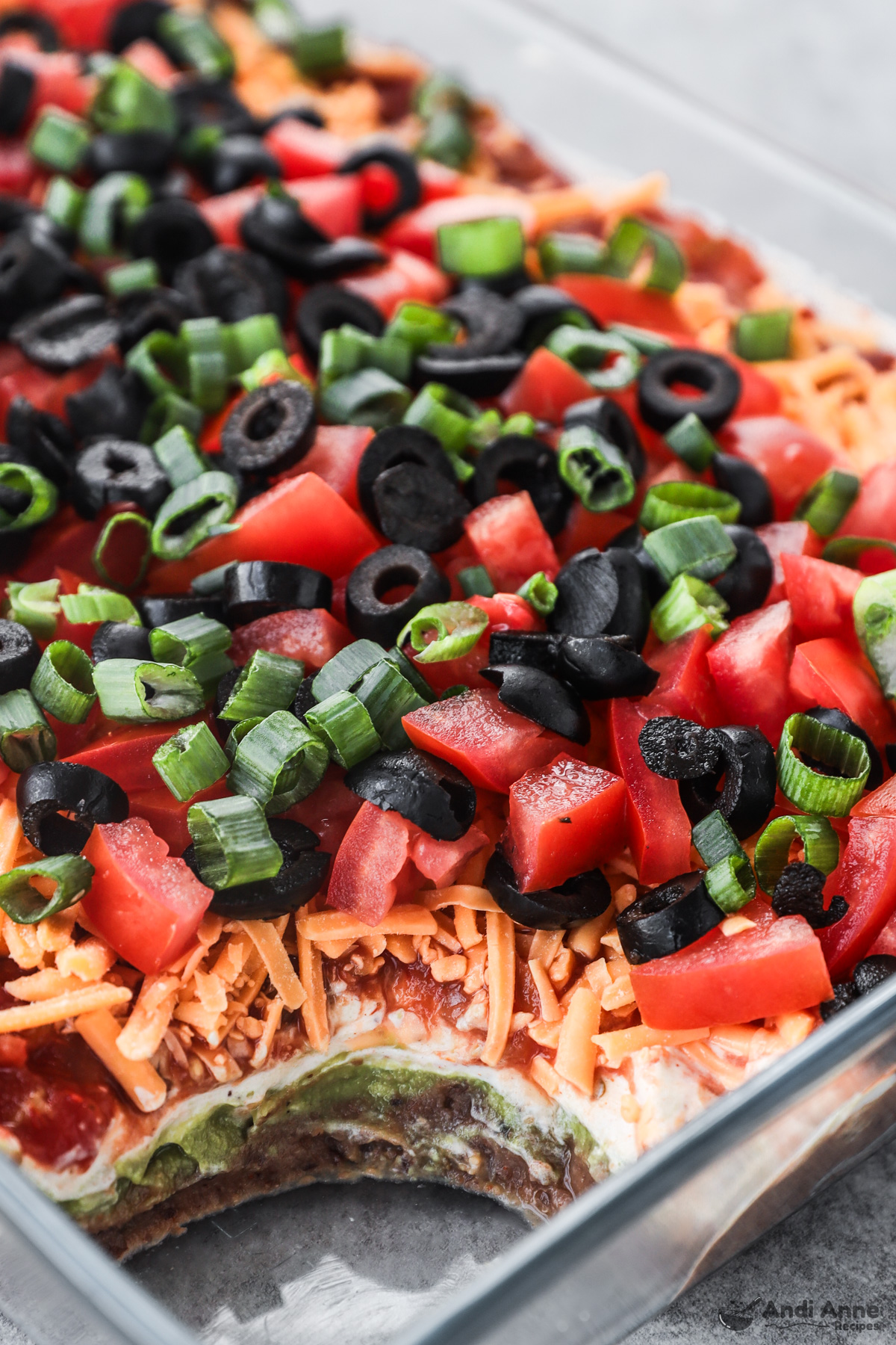 A casserole dish with 7 layer dip recipe including shredded cheese, sour cream, chopped tomatoes, black beans, and green onions.