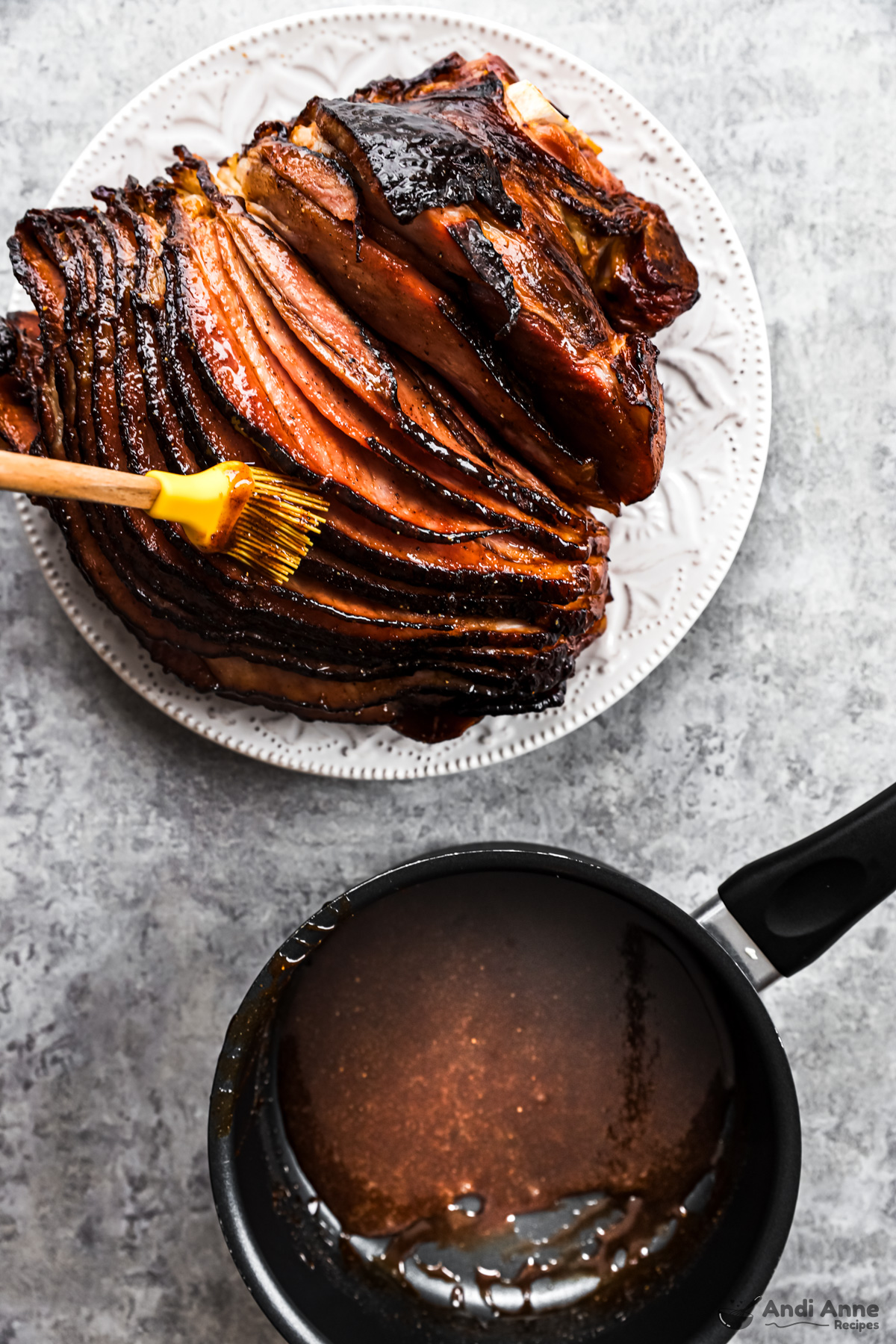 A ham on a plate with a brush adding glaze, plus a saucepan with glaze.