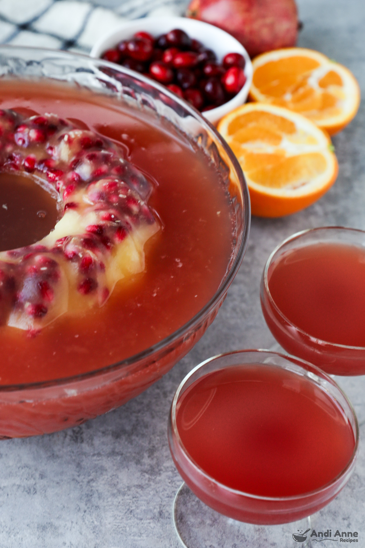 A punch bowl with red punch and large decorated ice cube with frozen pomegranate seeds, two glasses filled with punch, a sliced orange and a bowl of cranberries