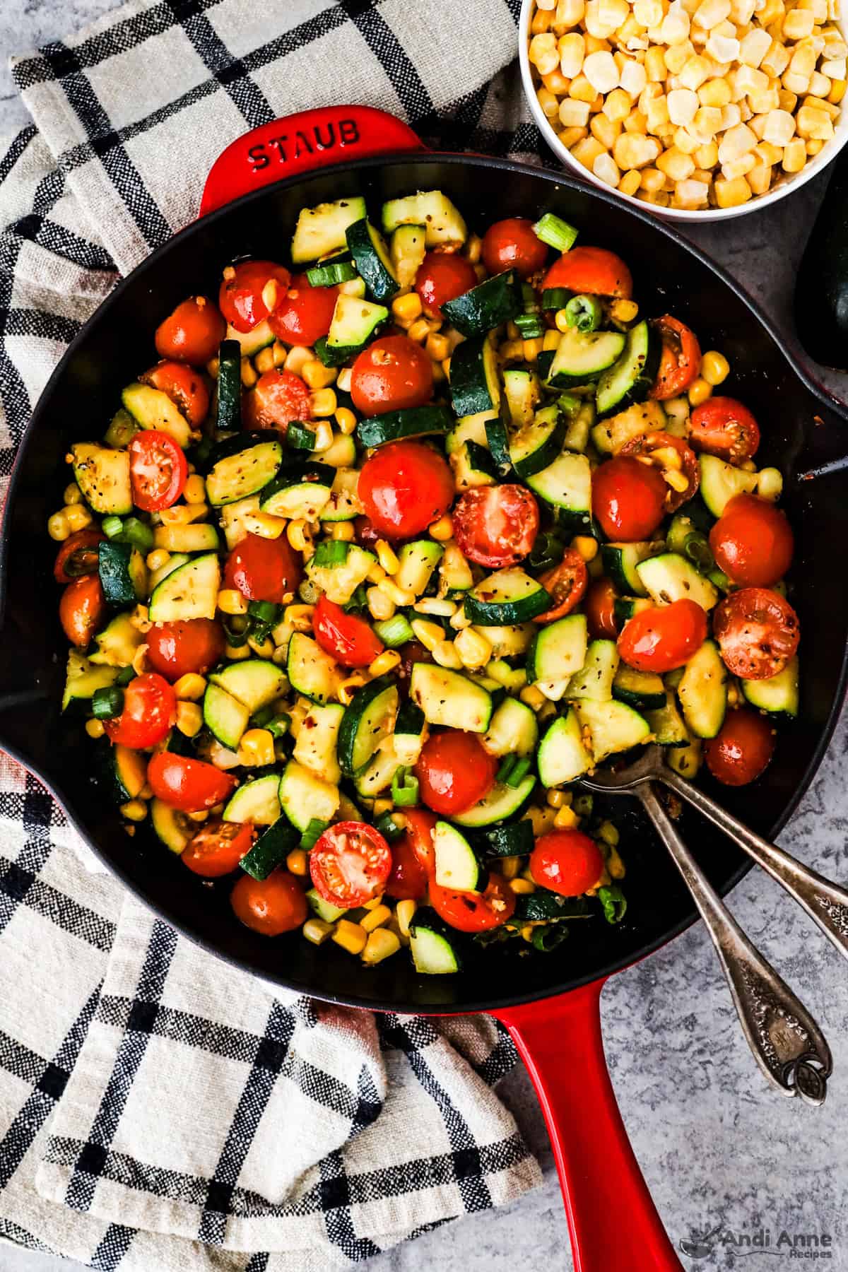 A skillet with chopped zucchini, tomatoes, corn and green onion and two spoons