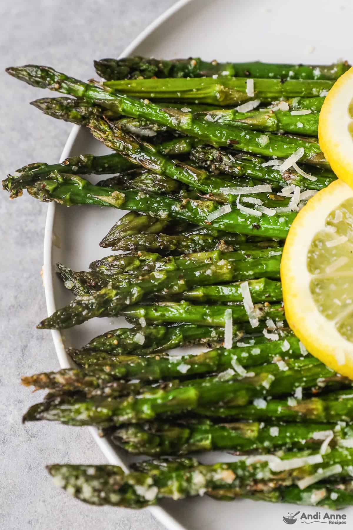 Closeup view of cooked asparagus on a white plate with grated parmesan cheese and two lemons. 